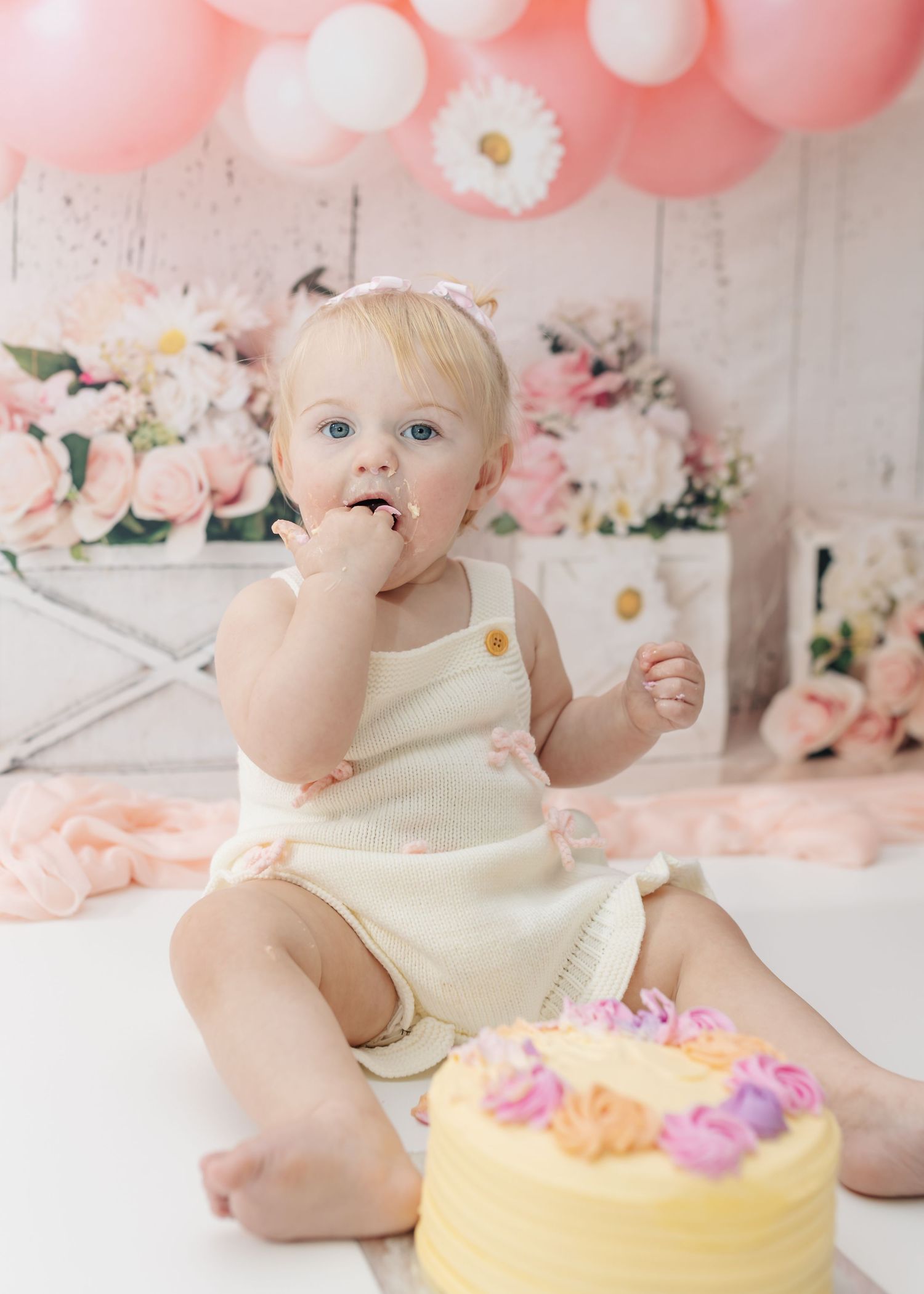 Baby explores yellow birthday cake during cake smash photo session with pink balloon backdrop.