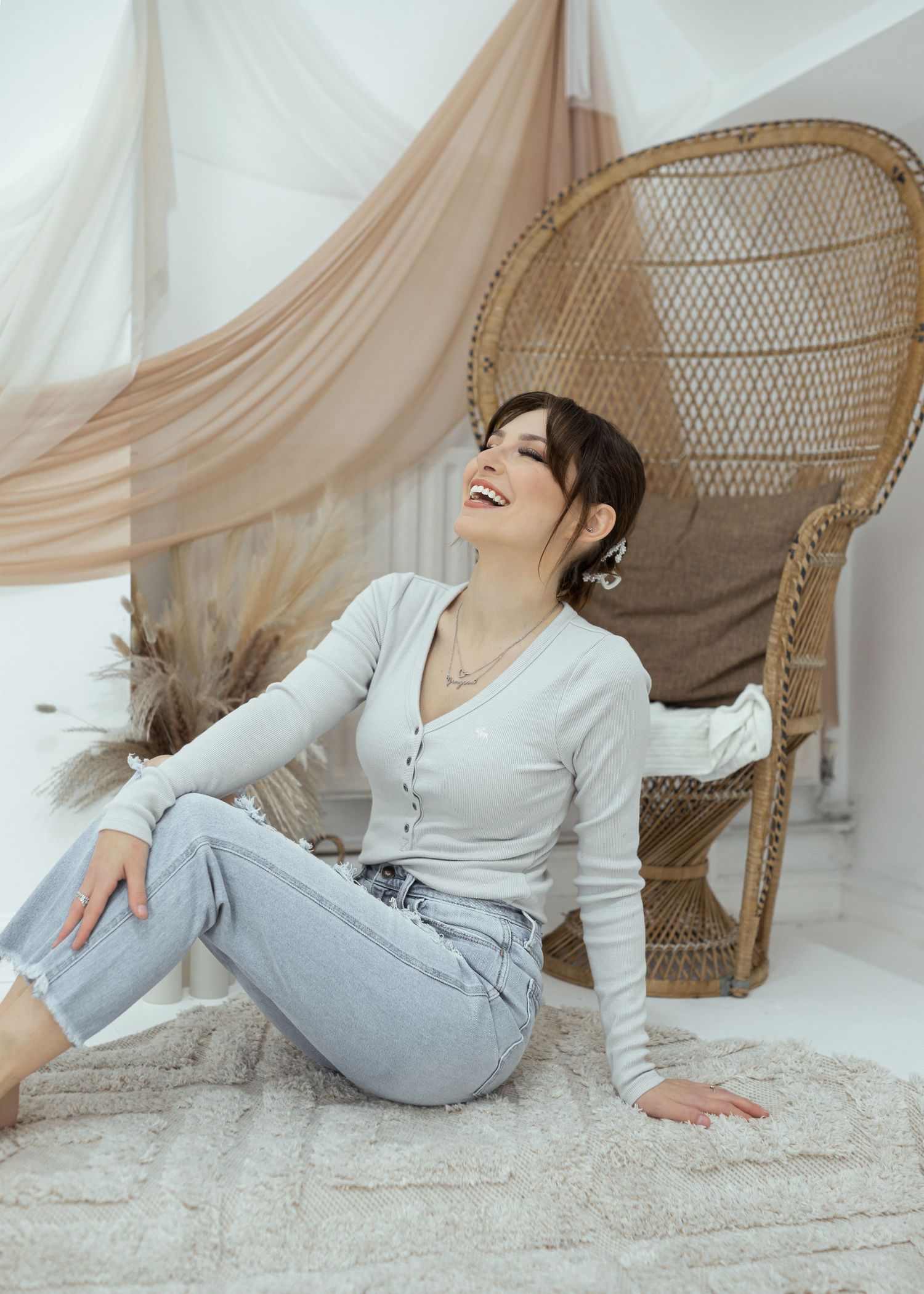 A person in comfortable attire poses playfully on a light carpet beside a peacock chair.