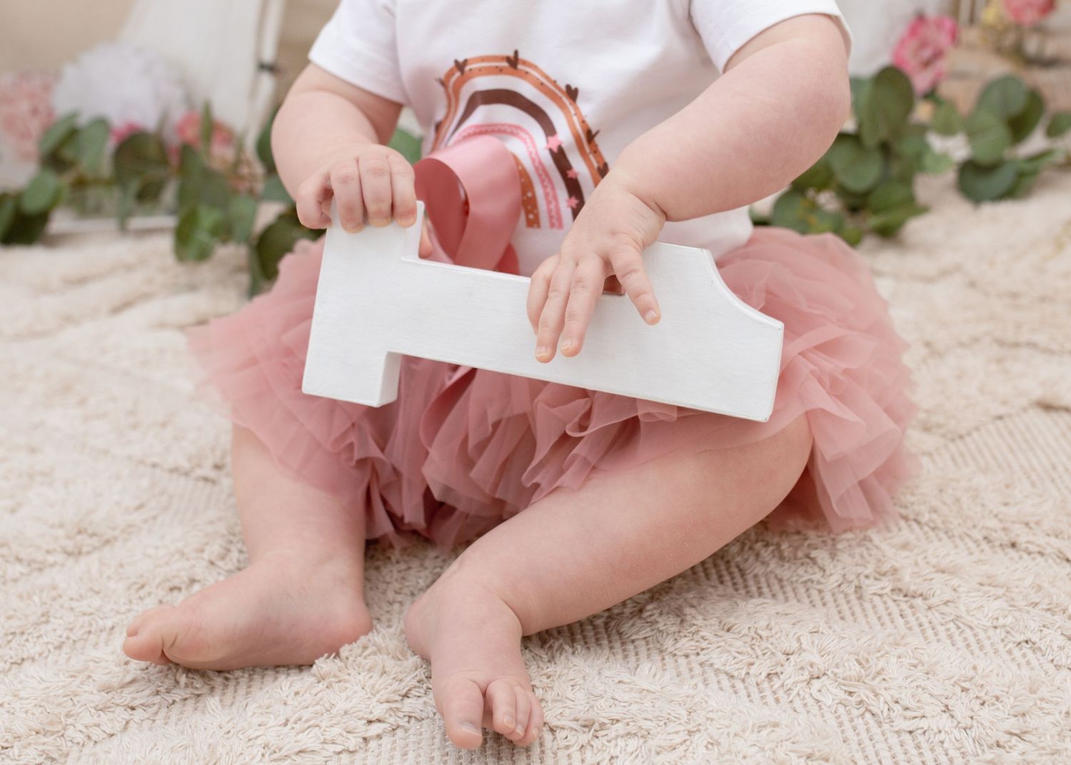 Small child in pink tutu holding white number prop while sitting.