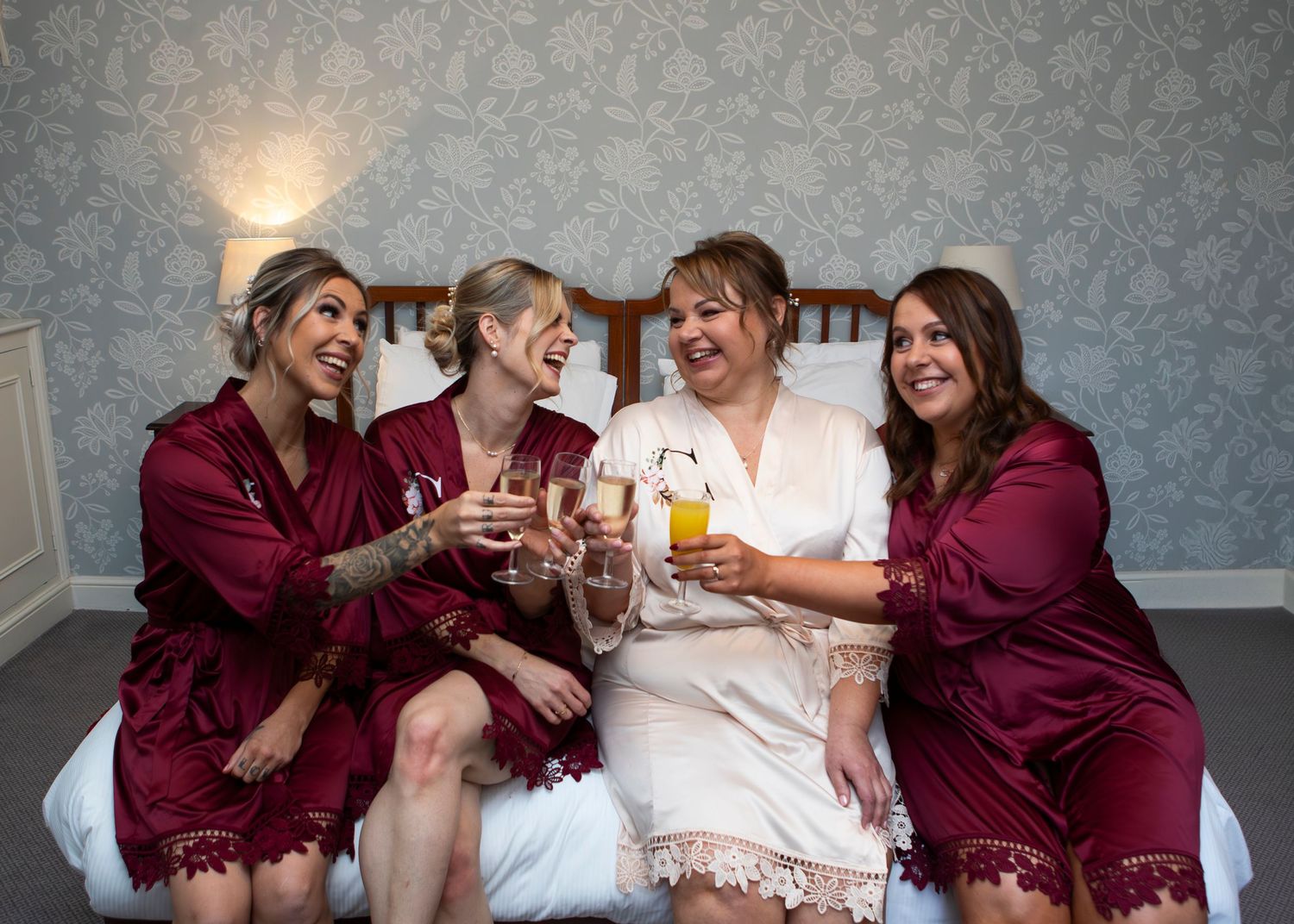 Bridesmaids in burgundy robes celebrating with drinks on a bed.