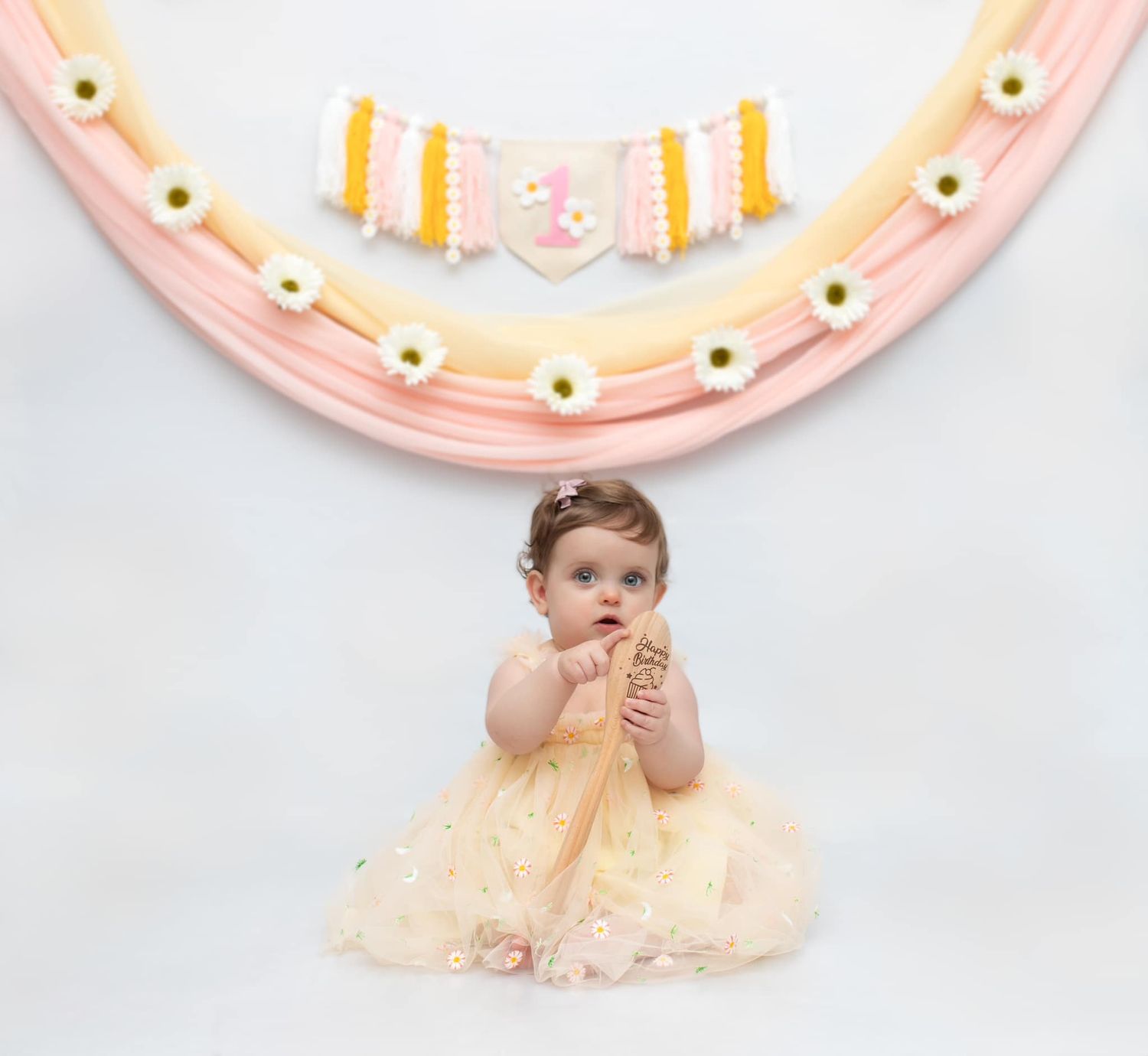 Baby in cream-colored dress sitting against pastel pink and yellow decorated backdrop.