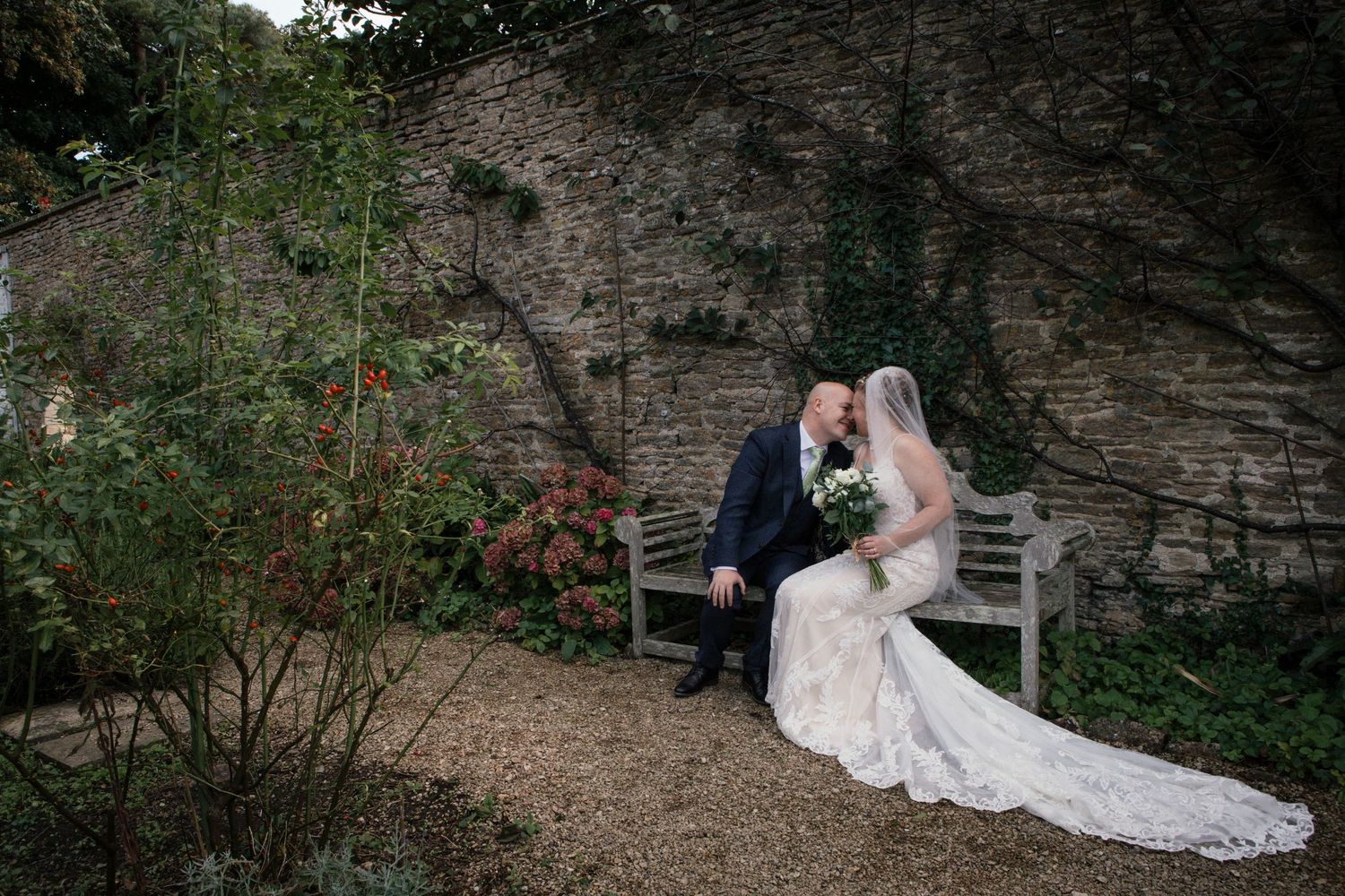 Romantic wedding couple poses against stone wall backdrop in garden setting.