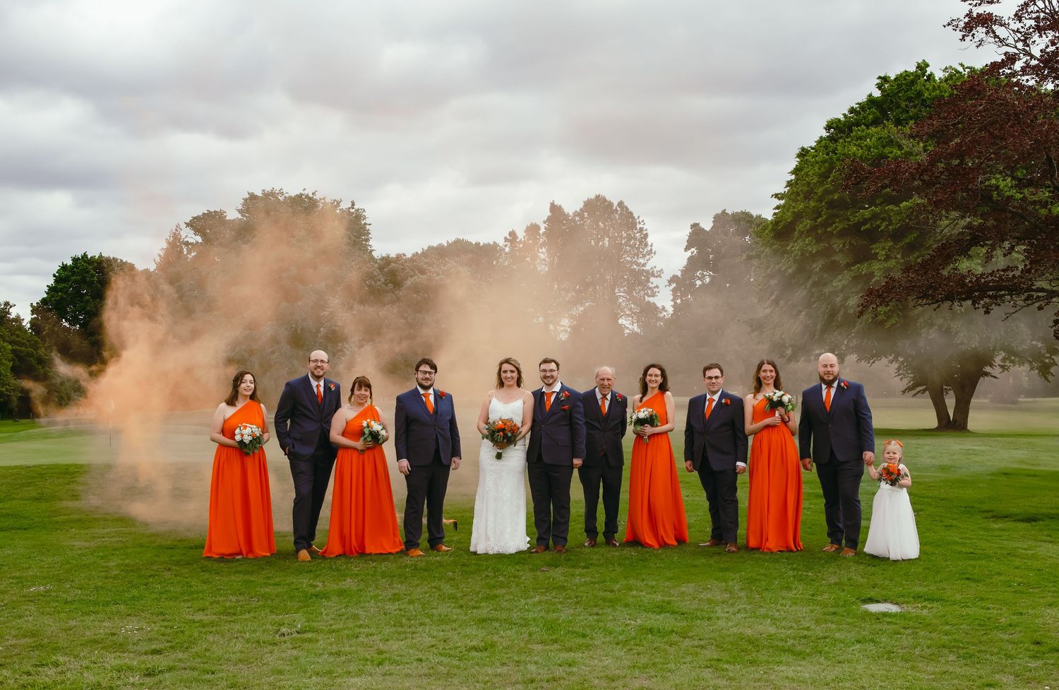 Wedding party poses on grass with orange smoke bombs creating a dramatic effect behind them.