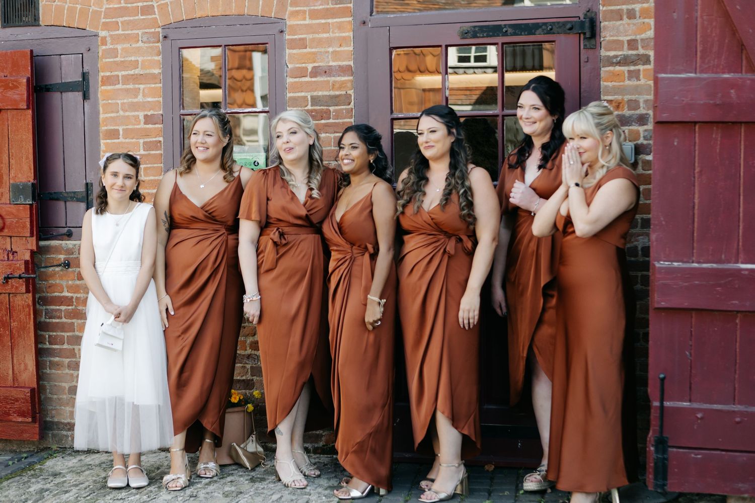 Row of bridesmaids in rust-colored dresses posing against brick wall.