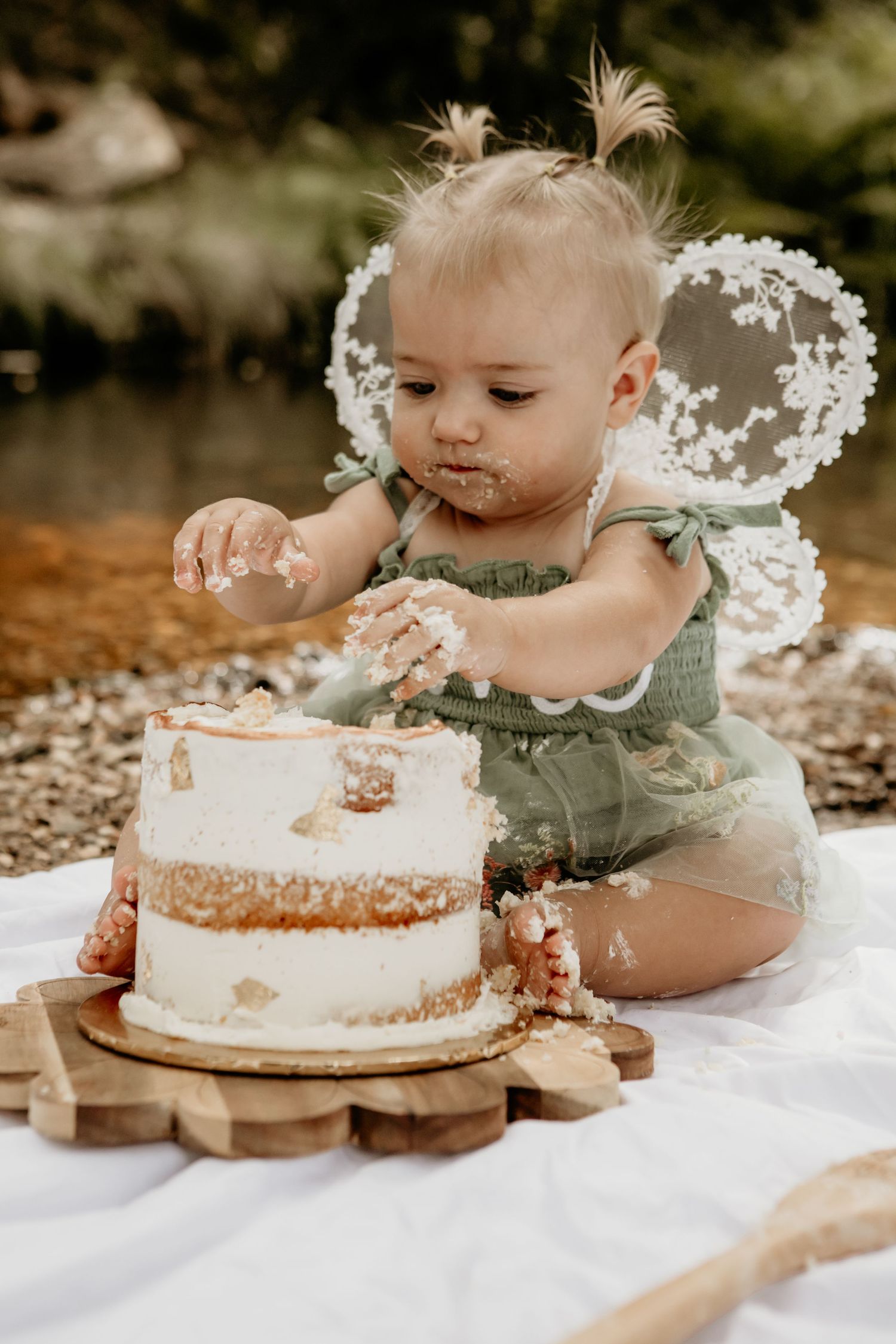 Small hands reaching for a white birthday cake decorated with flowers on a white blanket outdoors.