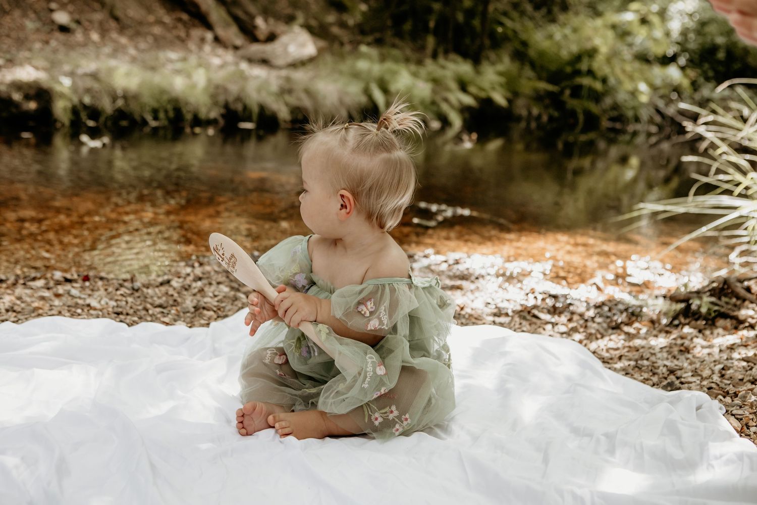 Baby in sage green romper sitting on white blanket outdoors for photoshoot.