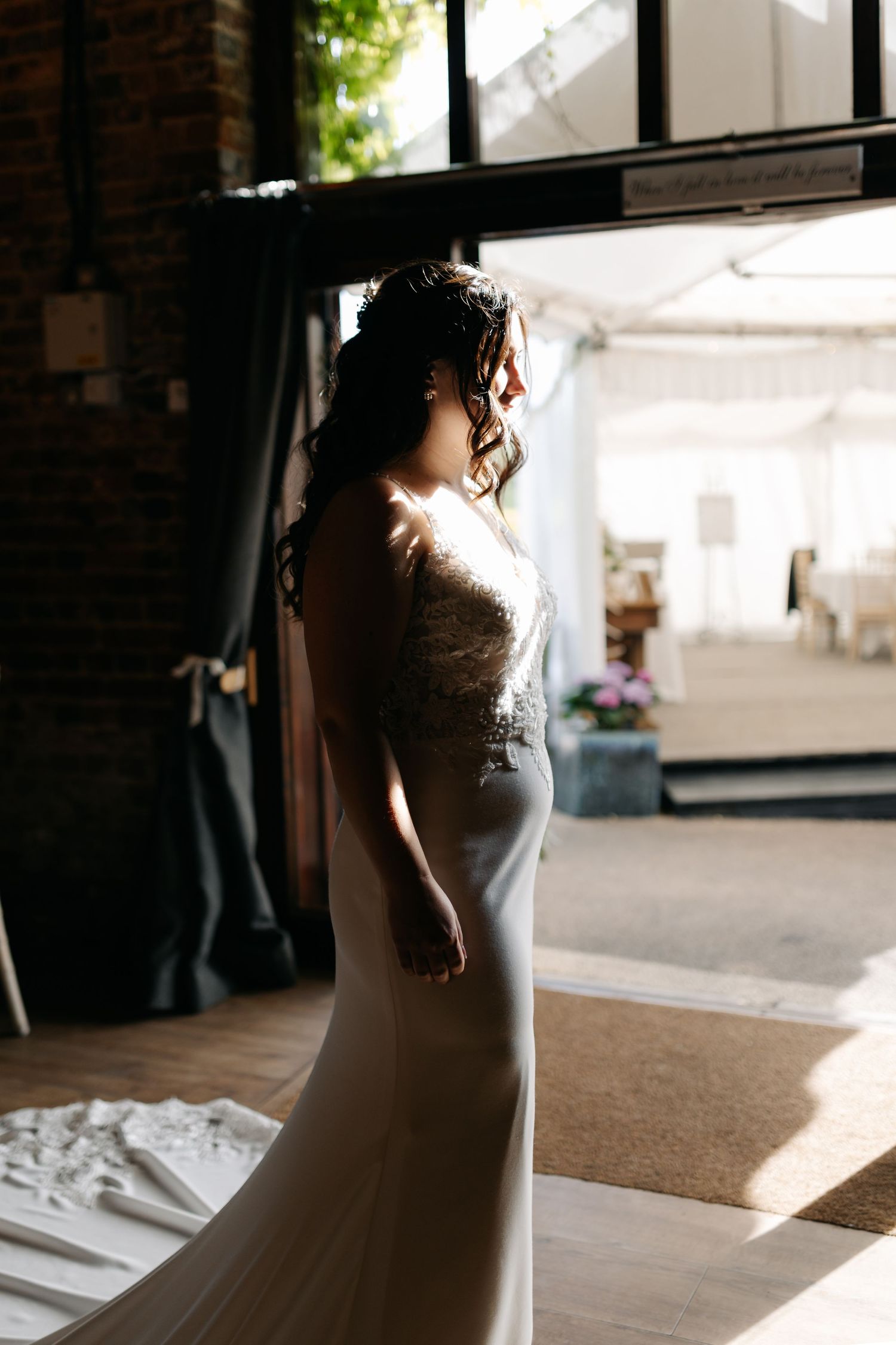 Silhouette of a bride in a lace wedding dress standing by a window in dramatic lighting.