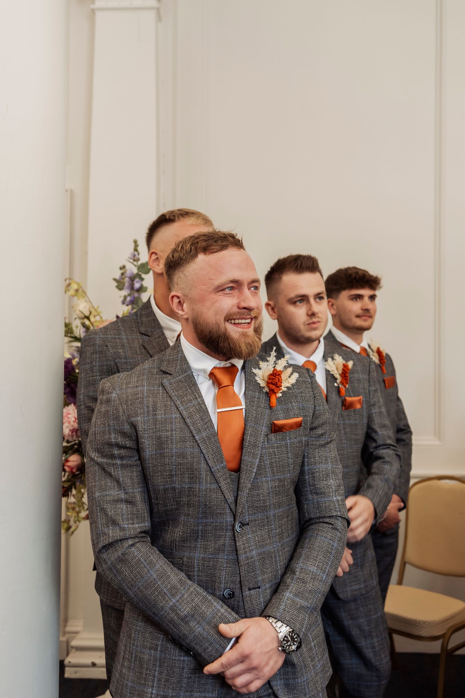Groomsmen in grey plaid suits with orange ties share a laugh during wedding ceremony.