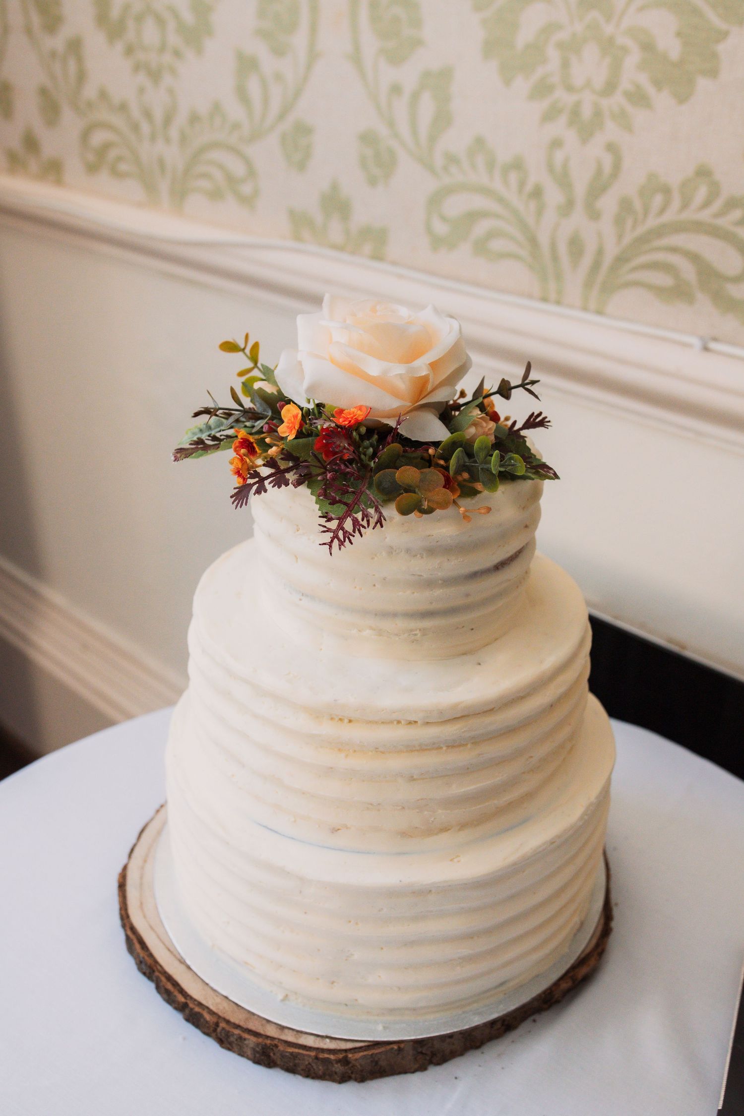 White wedding cake with textured frosting sits on rustic wood slice decorated with autumn florals.
