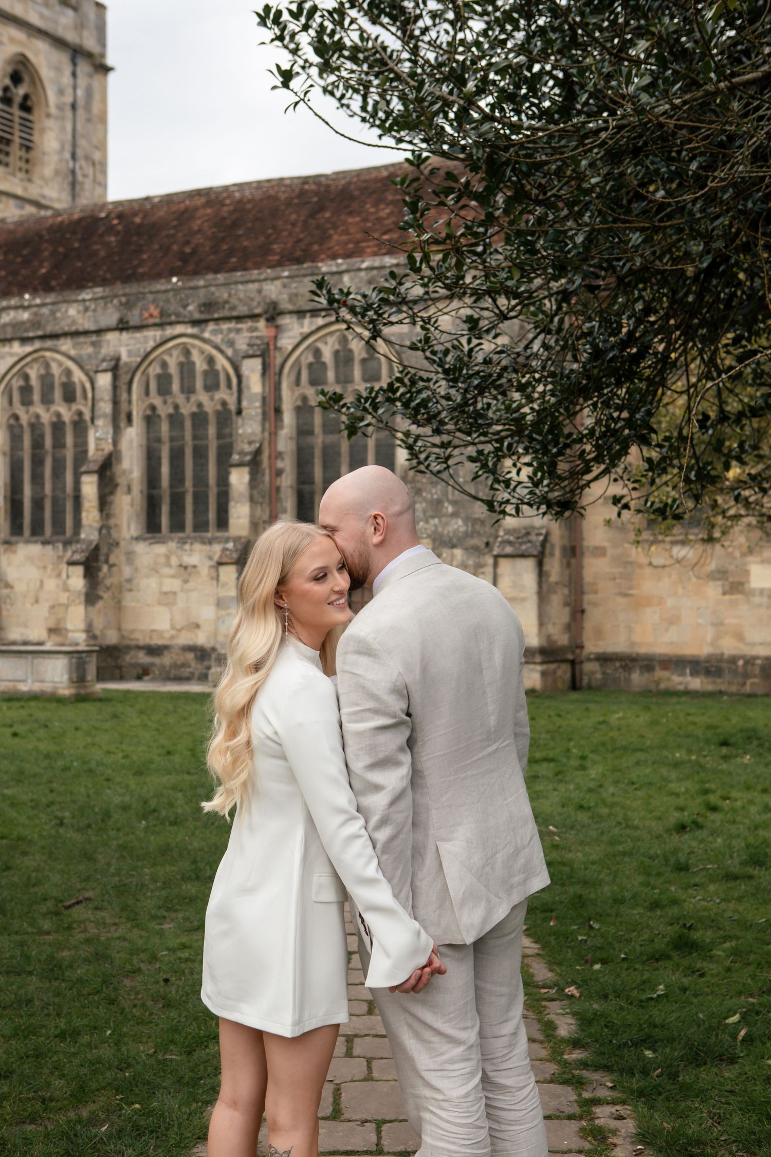 Couple in white and light gray outfits pose intimately outside historic stone church building.