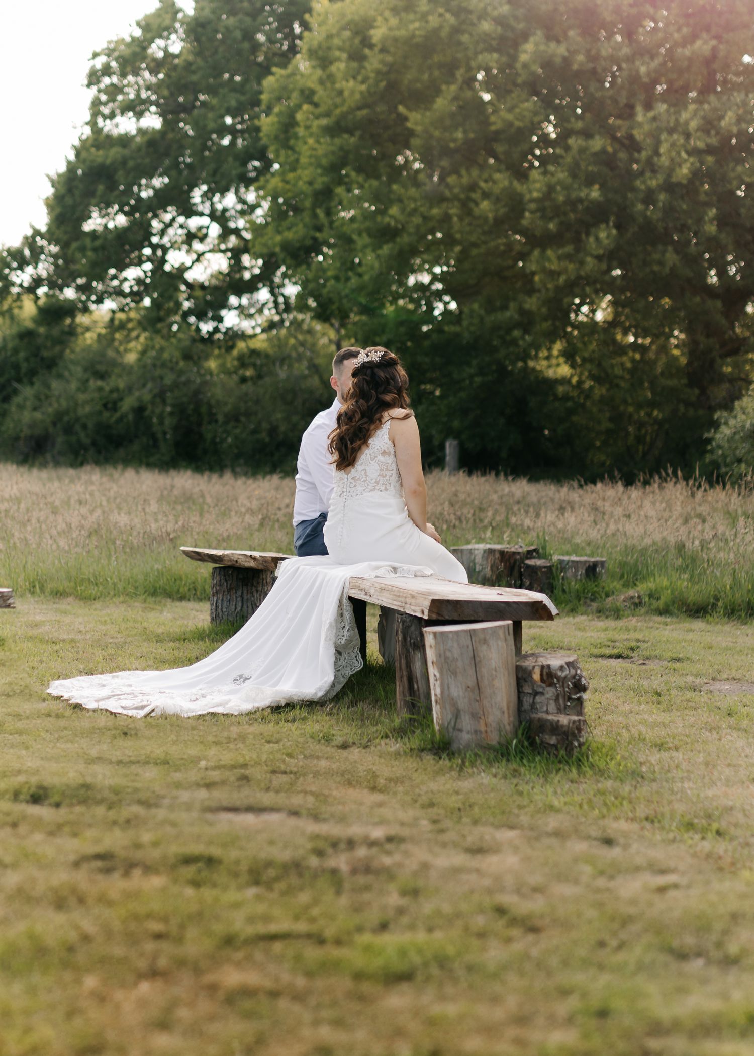 Couple sitting on rustic wooden bench in sunlit field during wedding photoshoot.