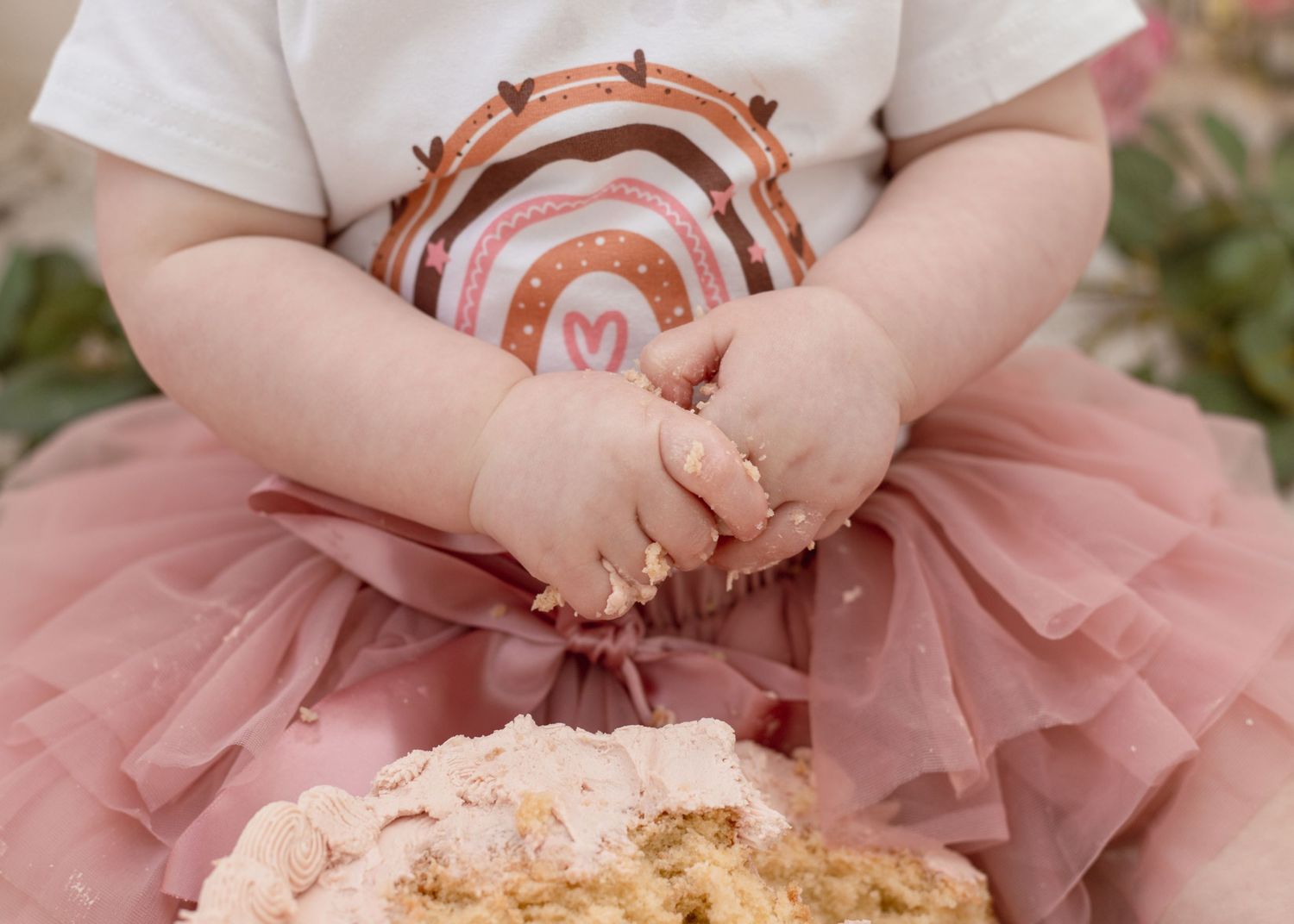 Baby in tulle skirt playing with crumbled cake on cream-colored blanket.