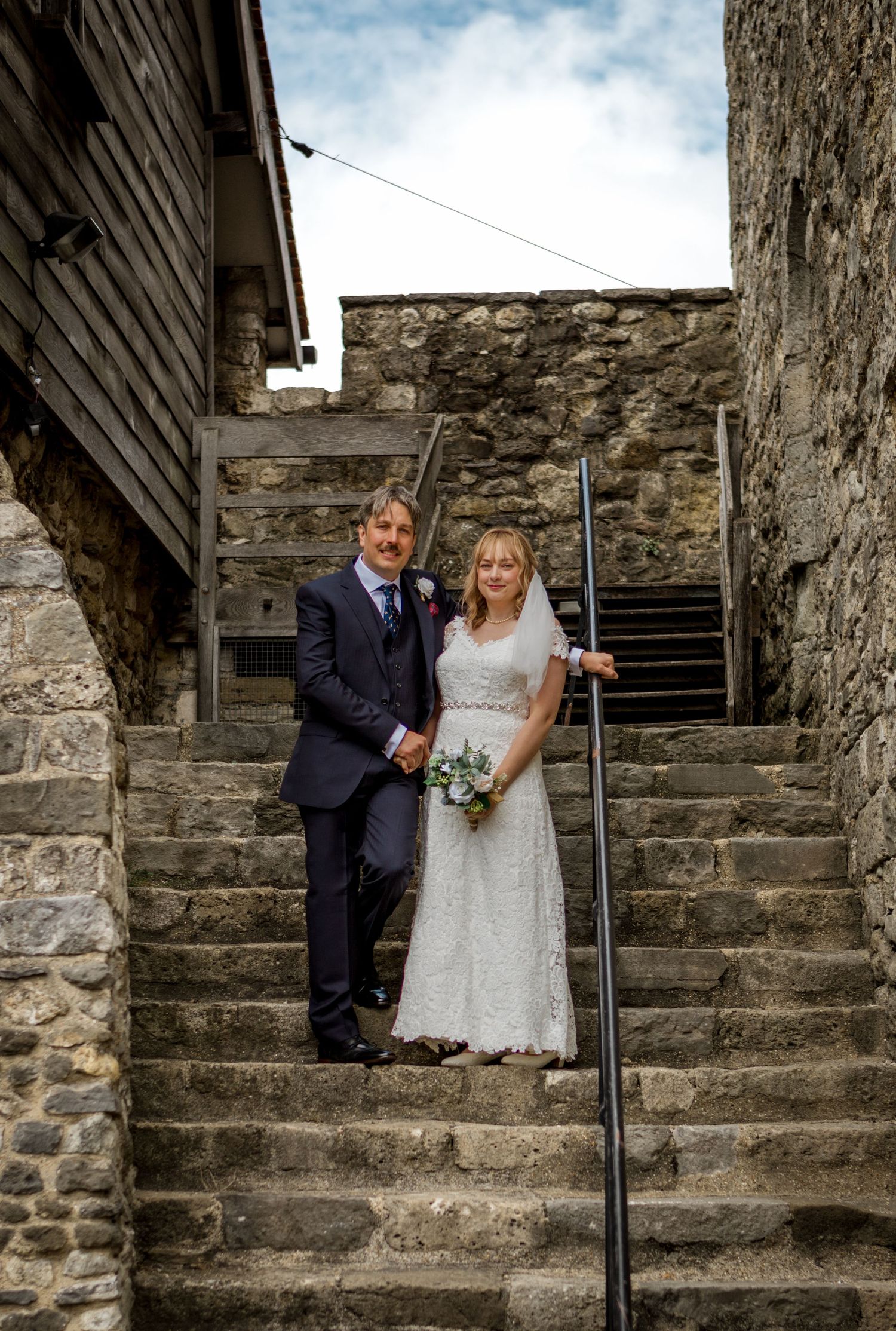 A couple stands on historic stone steps wearing wedding attire against an old stone wall backdrop.