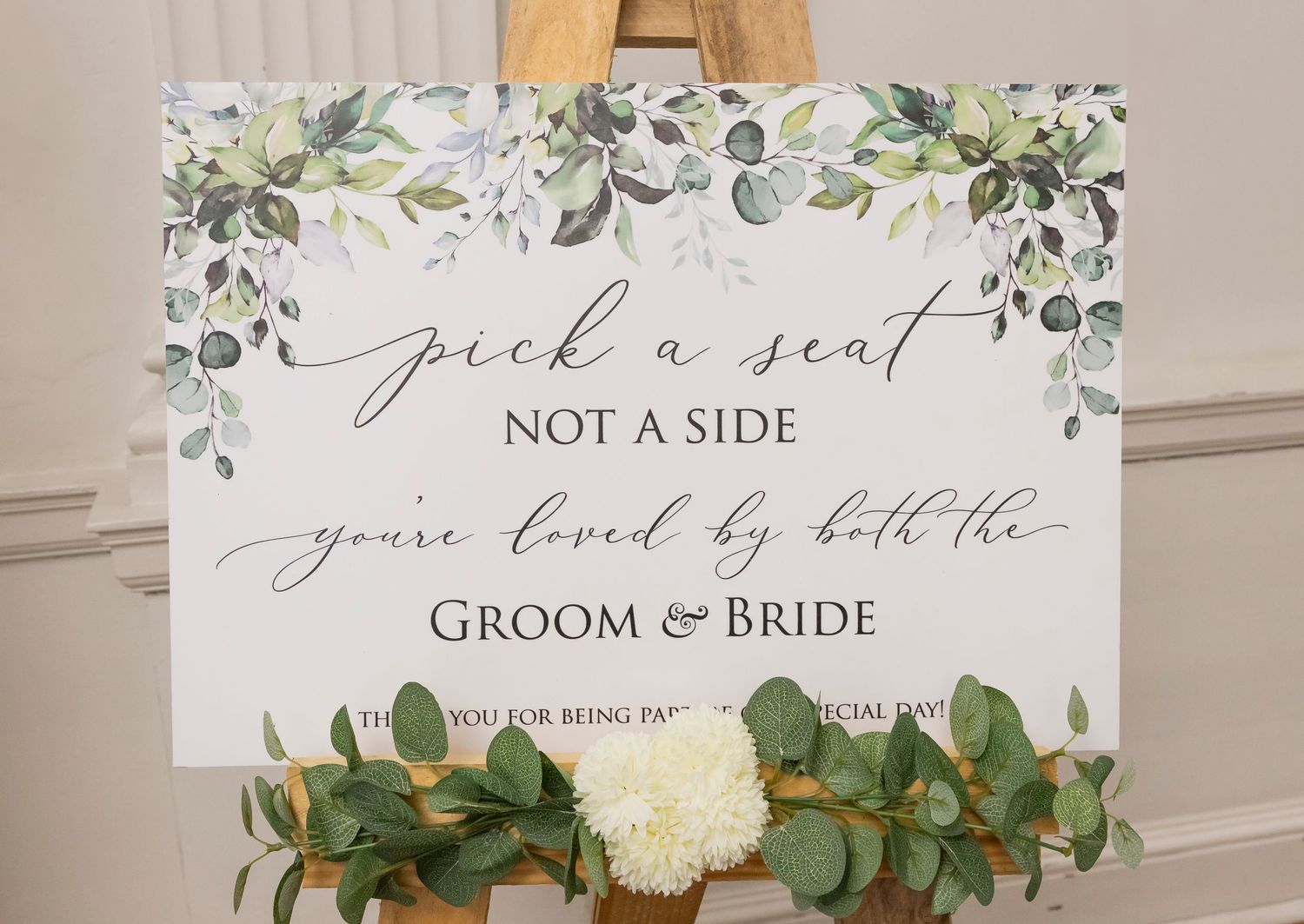 Elegant wedding signage with greenery and floral decorations against a neutral background.
