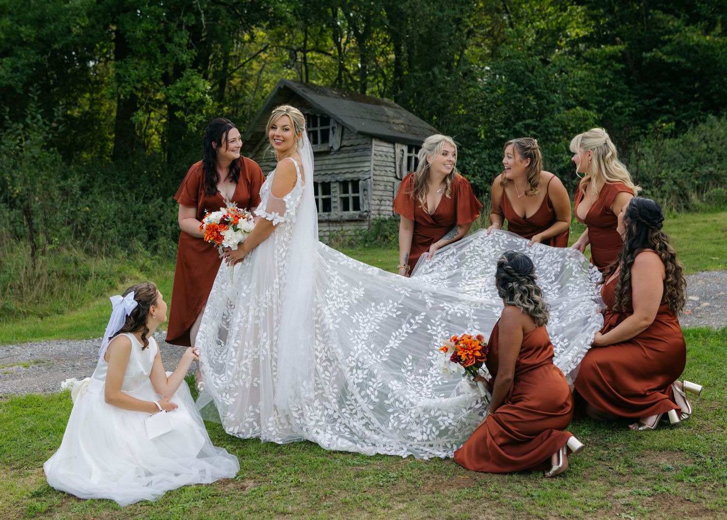 A bridal party in burgundy dresses arranges the bride's lace train on grass near a rustic cabin.