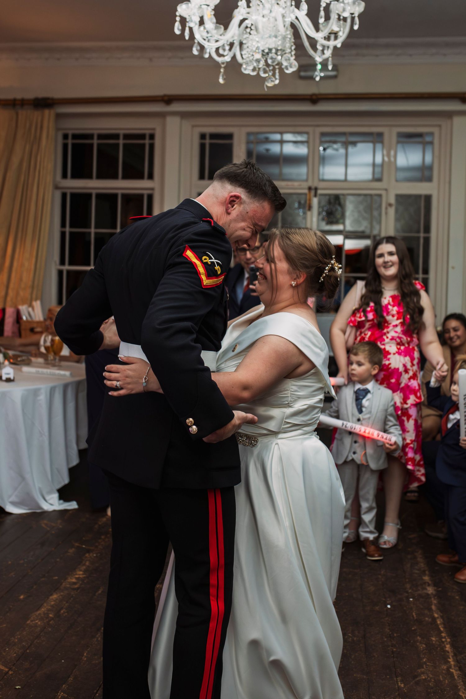 Wedding couple shares their first dance under crystal chandelier as guests watch.