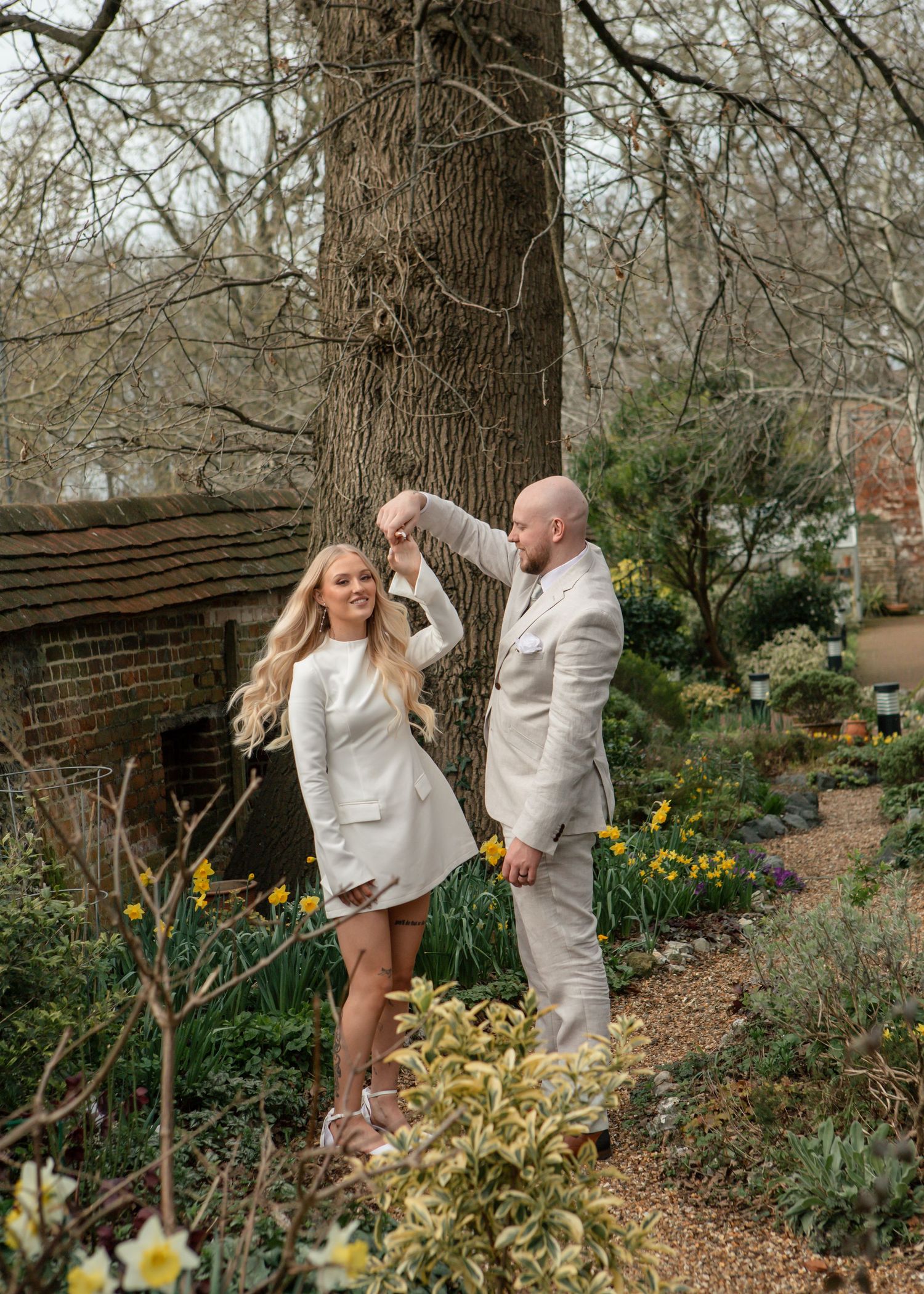 A couple in white outfits poses playfully in a garden setting with blooming daffodils and trees.