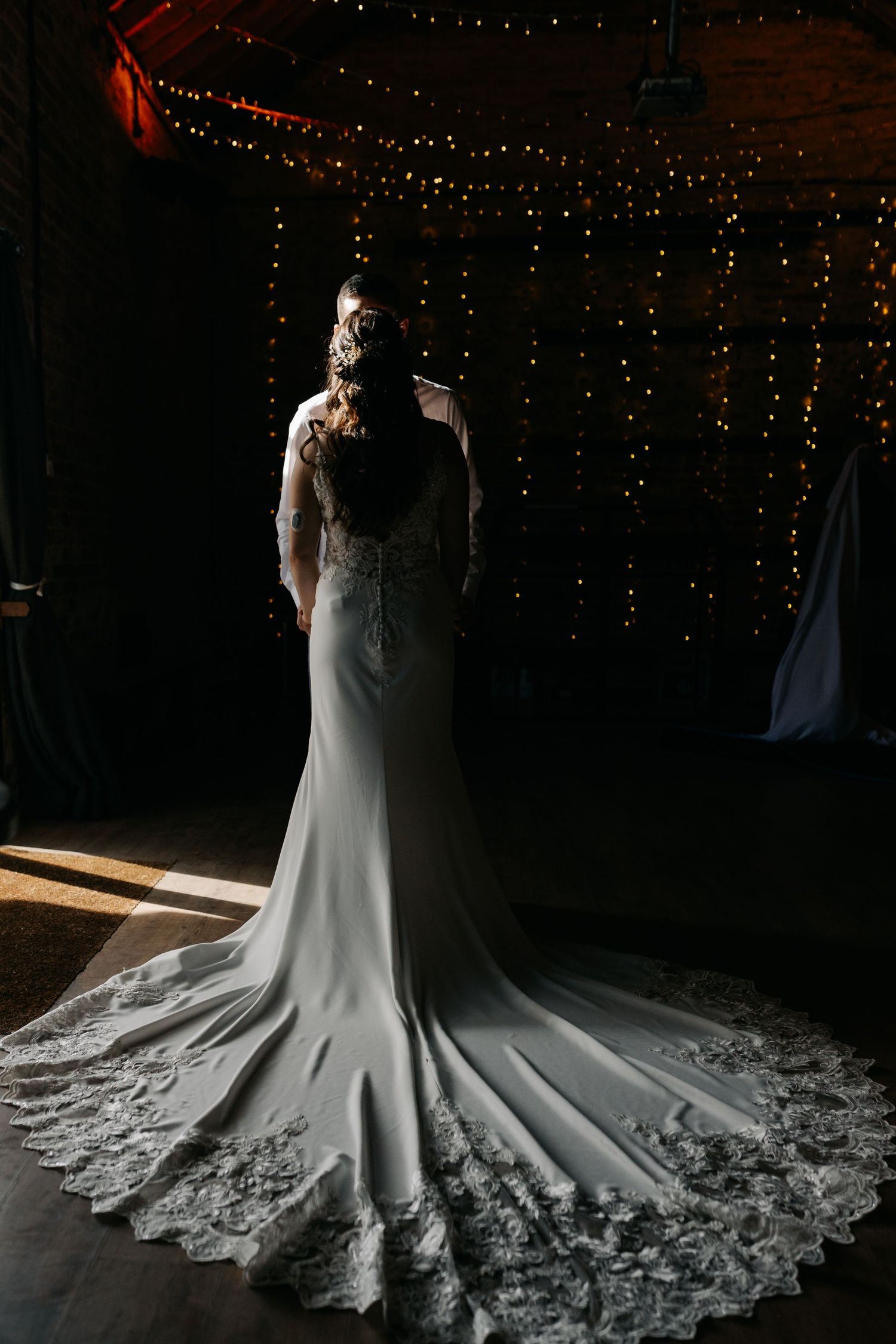 Wedding dress with intricate lace train displayed against dark background with twinkling lights.