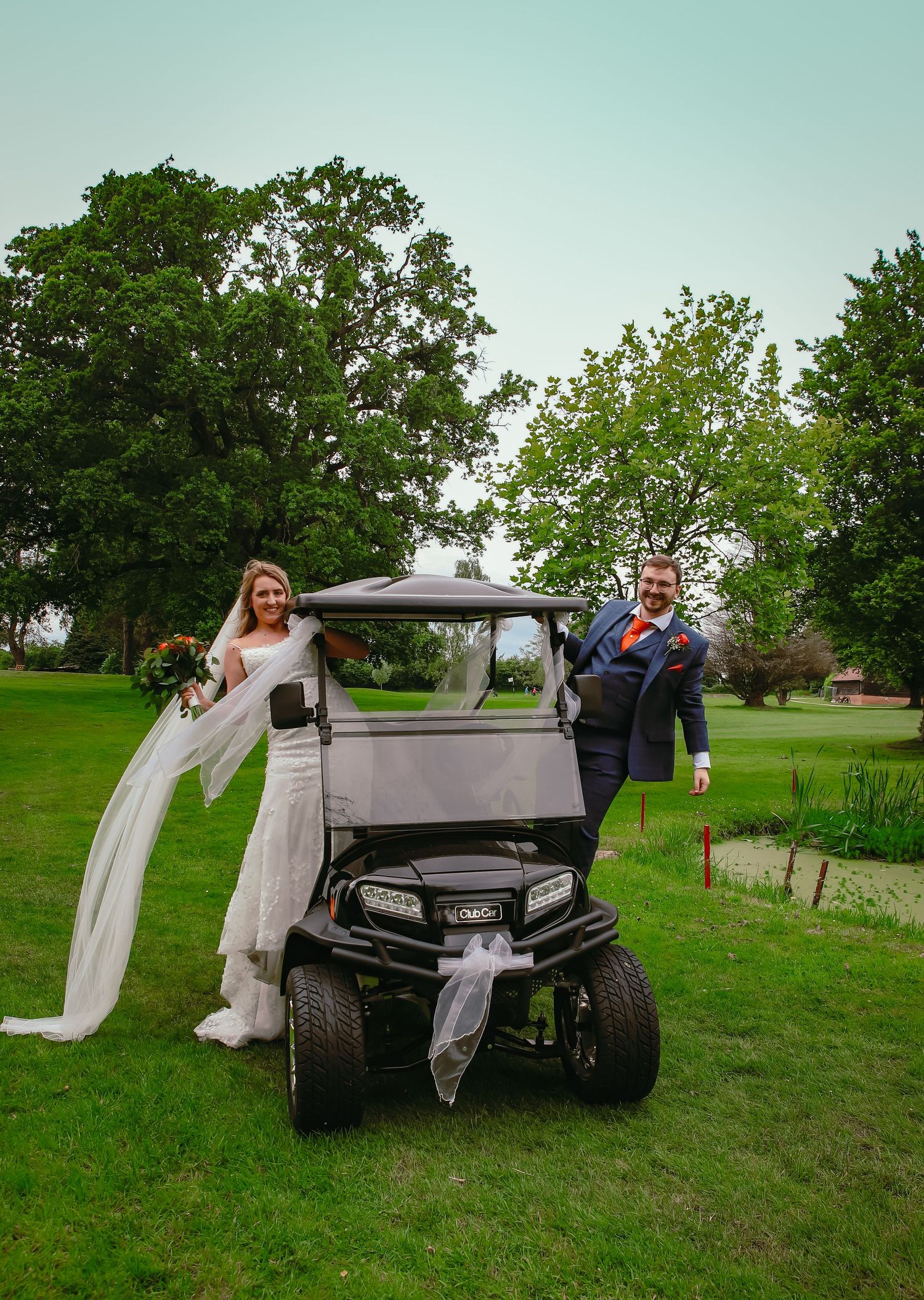 Newlyweds pose playfully with a golf cart on their wedding day in a scenic outdoor setting.