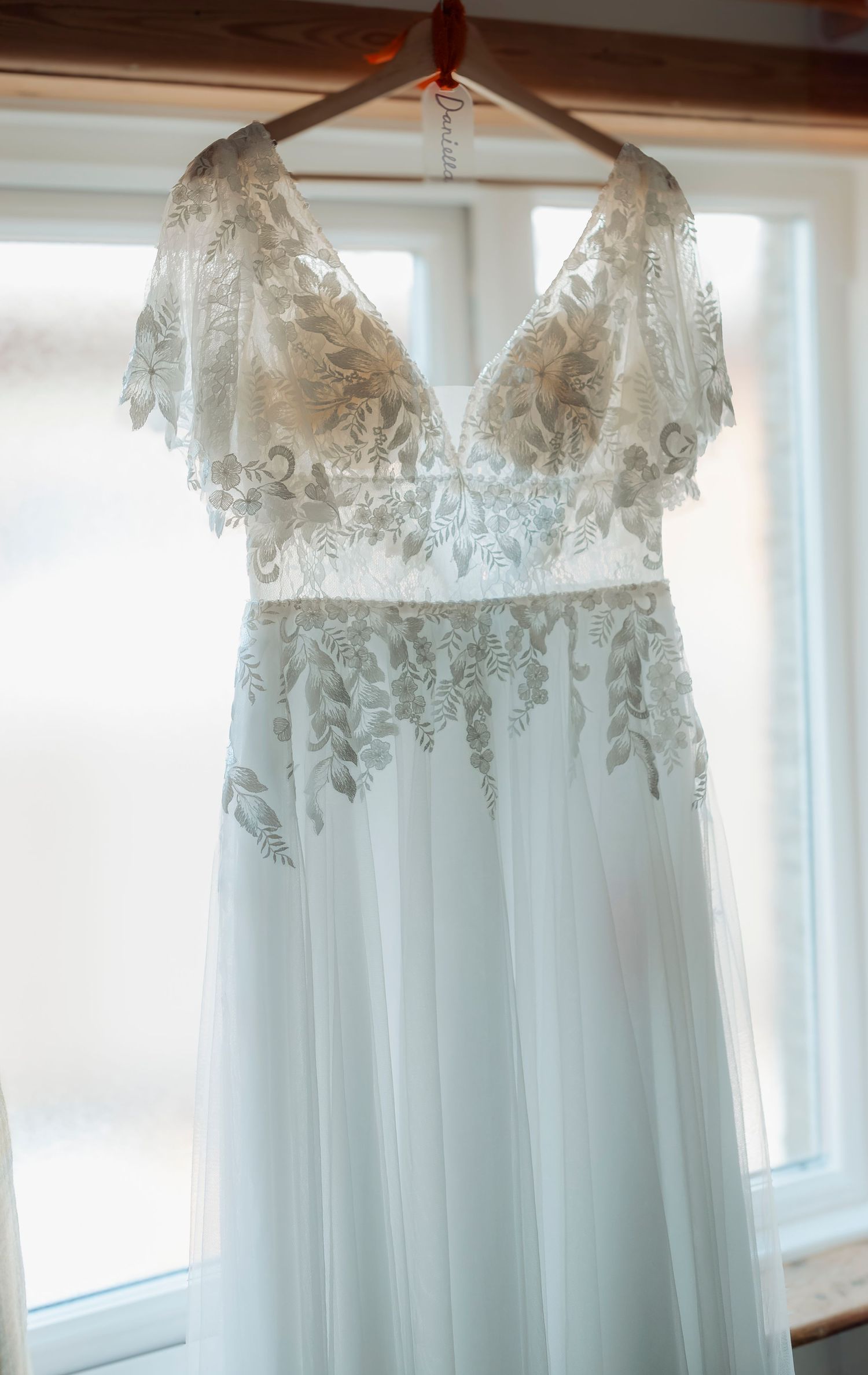 A delicate white wedding dress with lace details hanging in front of a window.