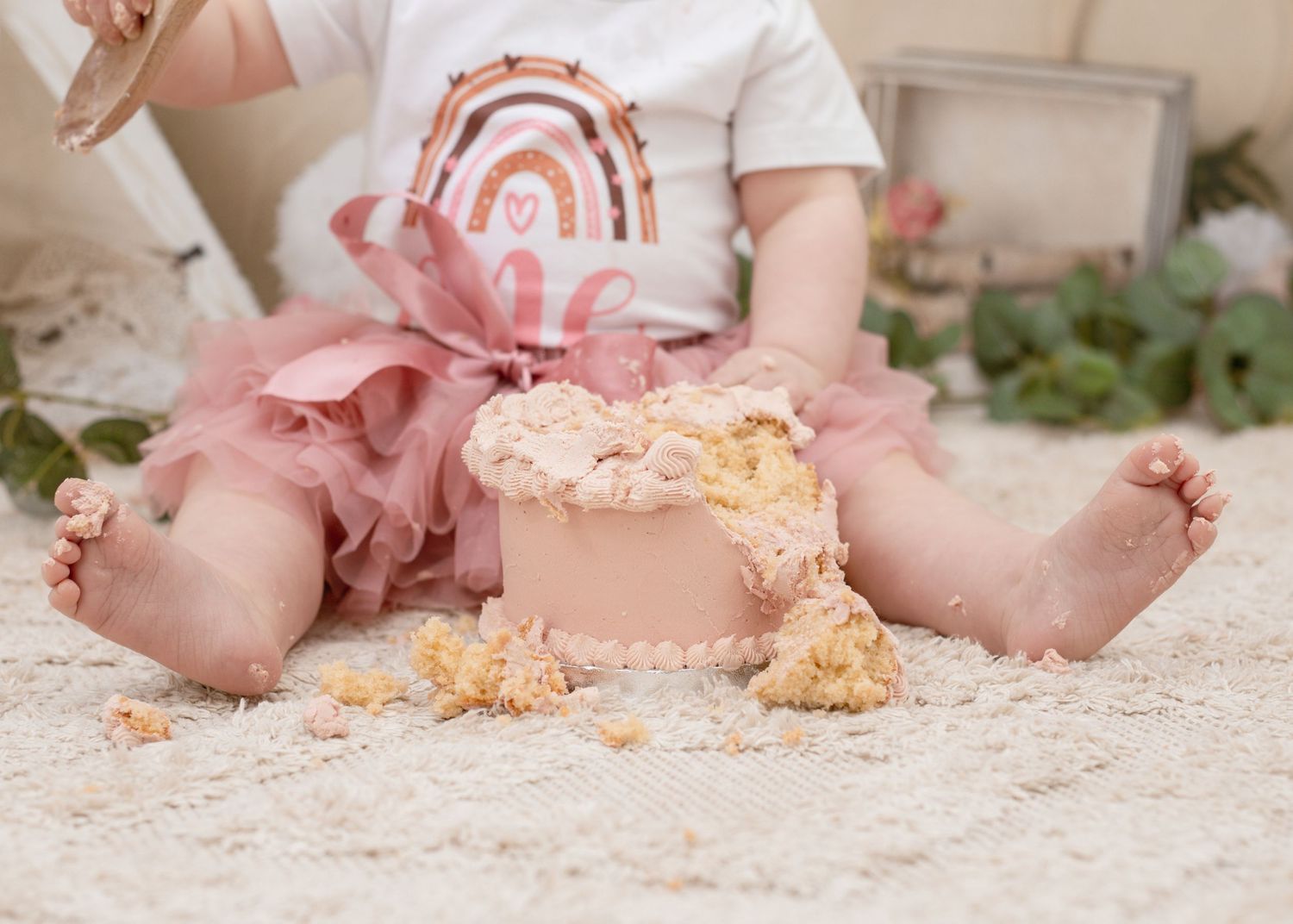 Baby in white shirt with pink tutu sitting on carpet with destroyed cake.