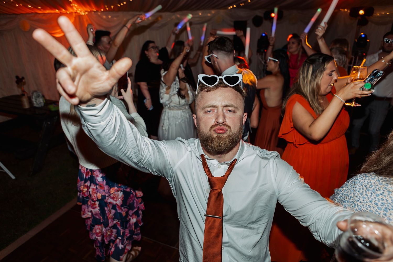 Party guests dancing and having fun at a wedding reception under orange lighting and draped ceiling.
