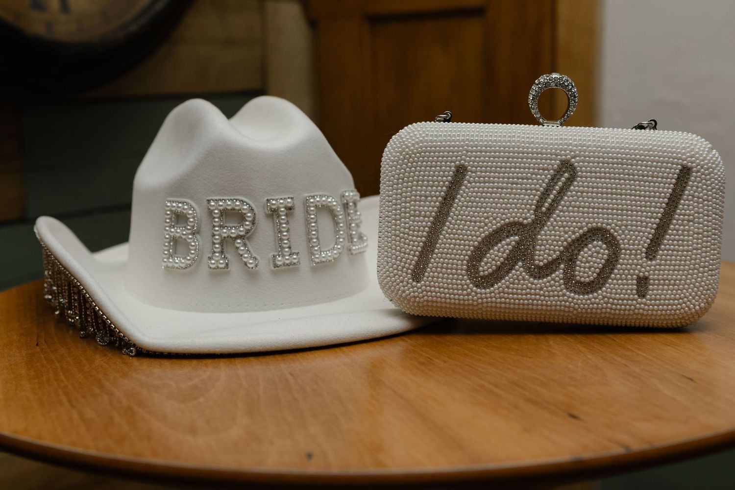 A white cowboy hat reading 'BRIDE' next to a clutch saying 'I do' on wooden surface.