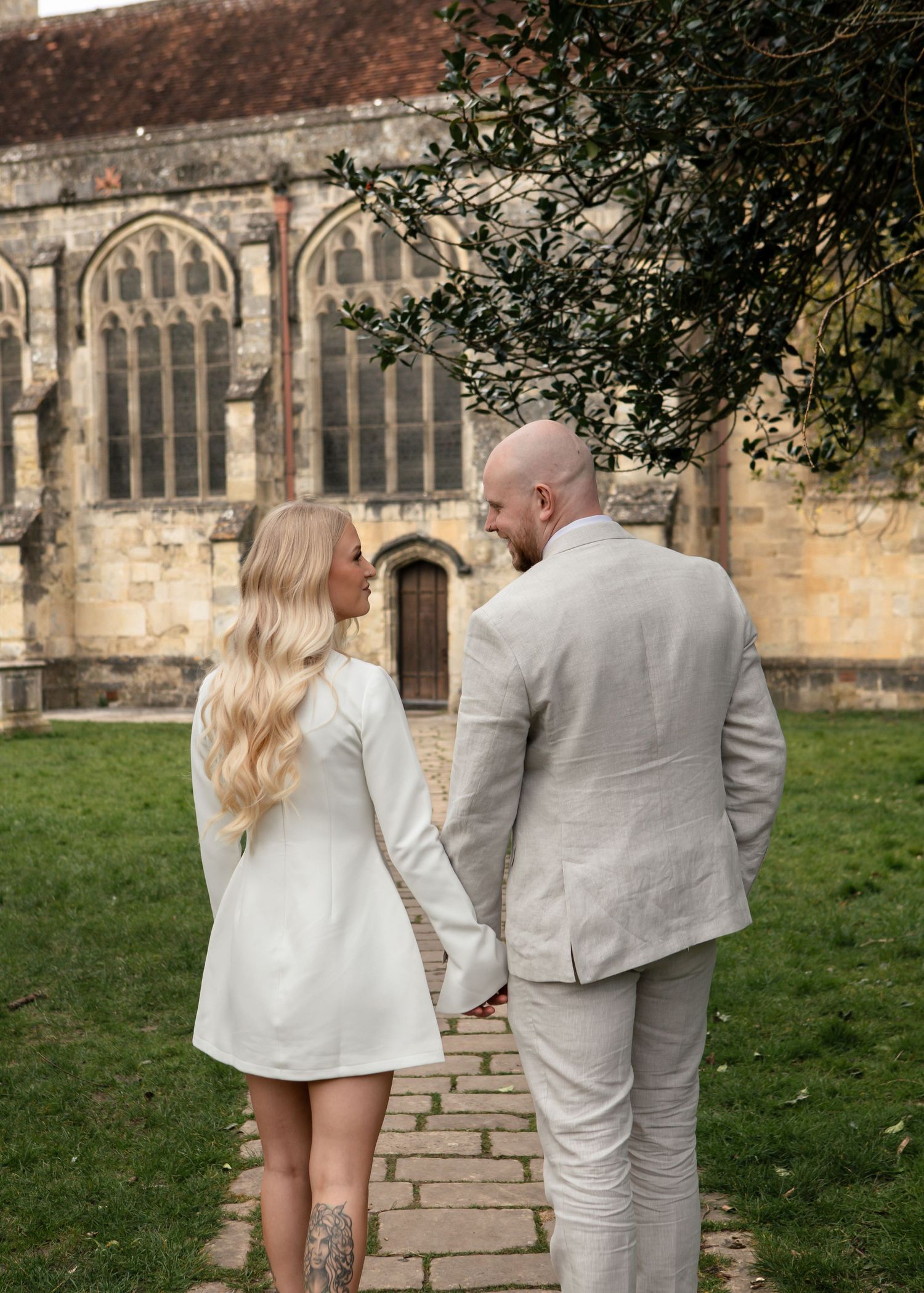 Couple in coordinating white and gray outfits walk along stone path by historic church.