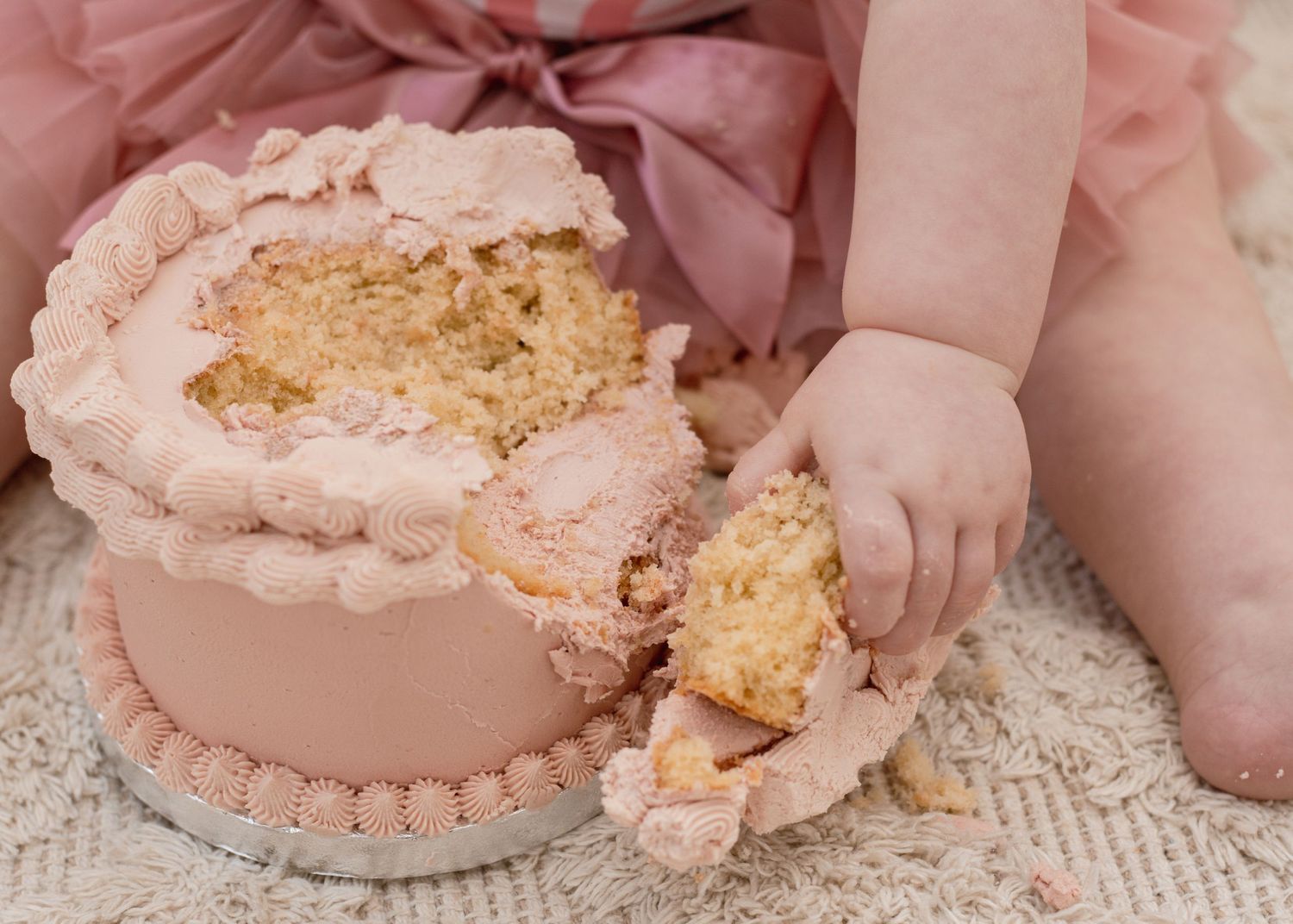 Pink frosted cake being smashed and eaten on a cream colored carpet.