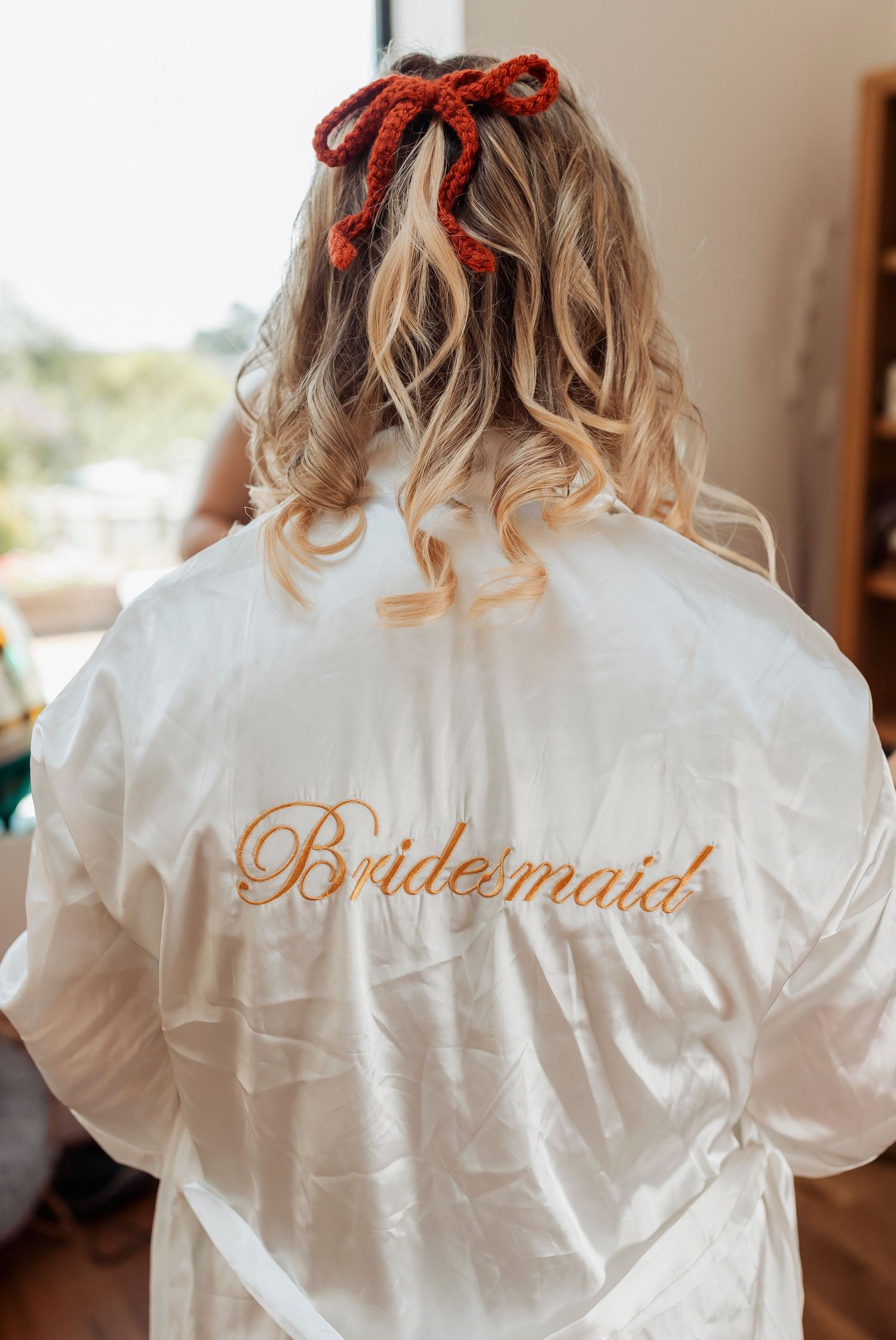 Back view of a white satin robe with 'Bridesmaid' embroidered in gold and a red bow hair accessory.