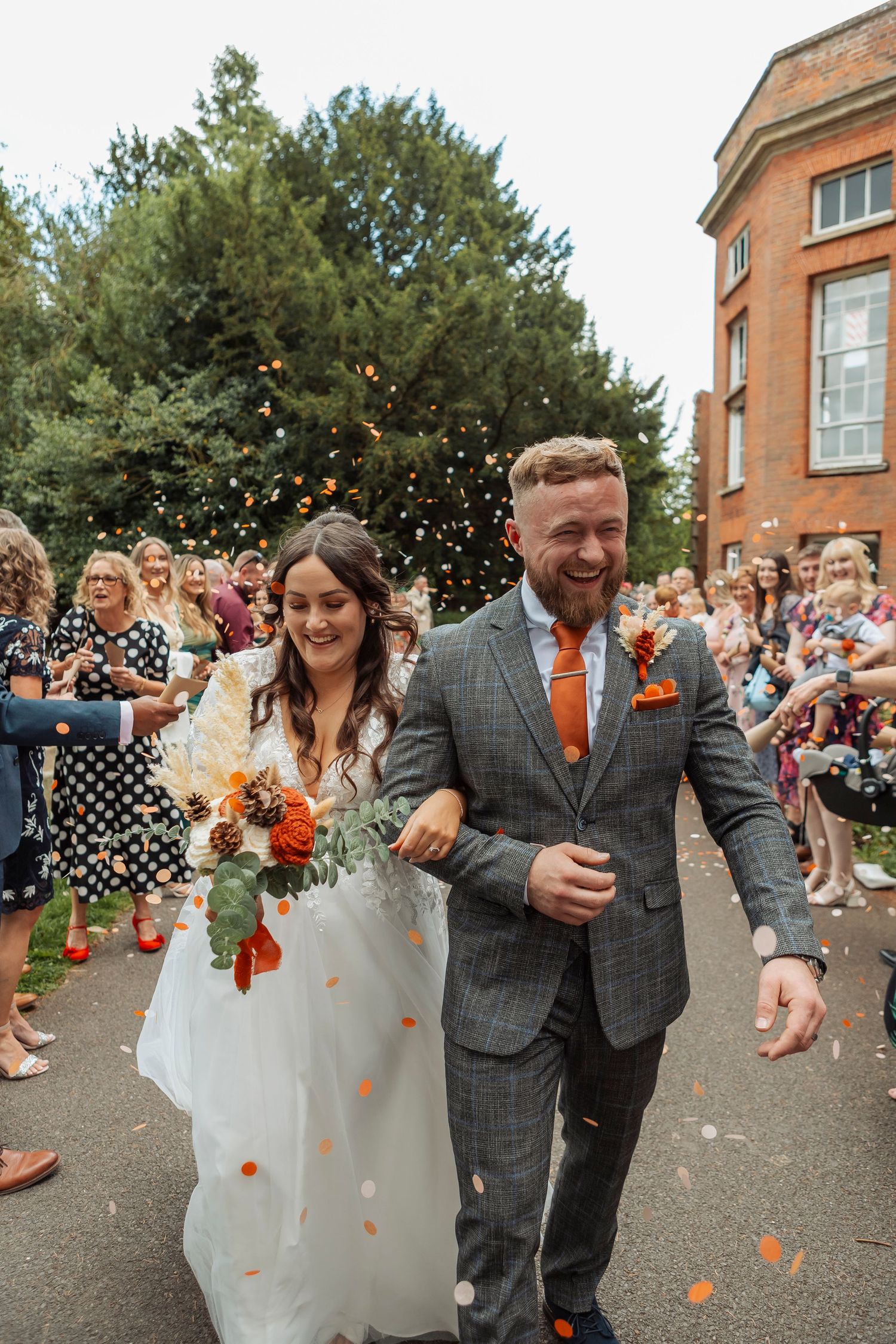 Happy couple runs through shower of confetti outside historic brick building during wedding exit.