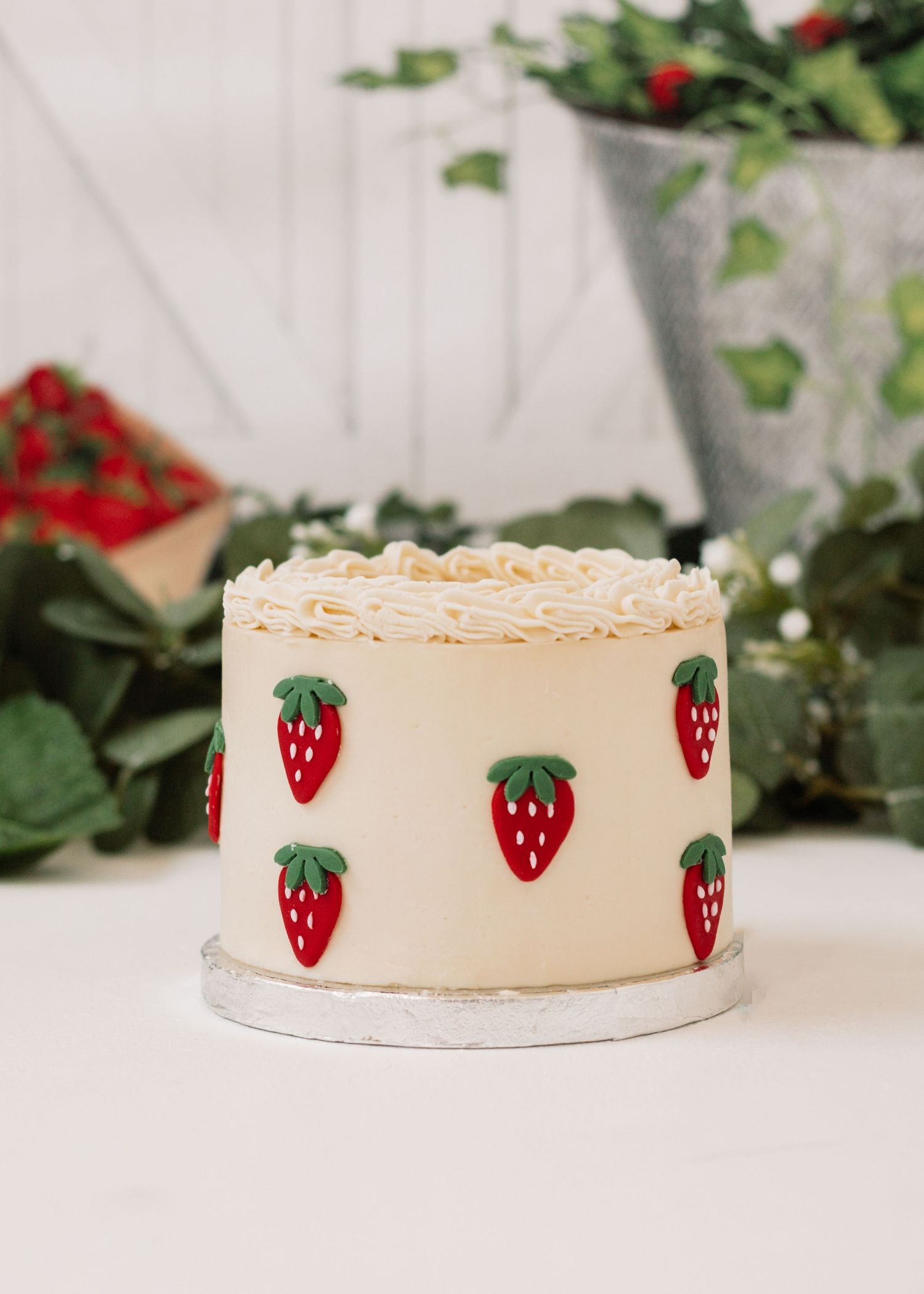 Cream colored cake decorated with cute strawberry designs against green foliage backdrop.