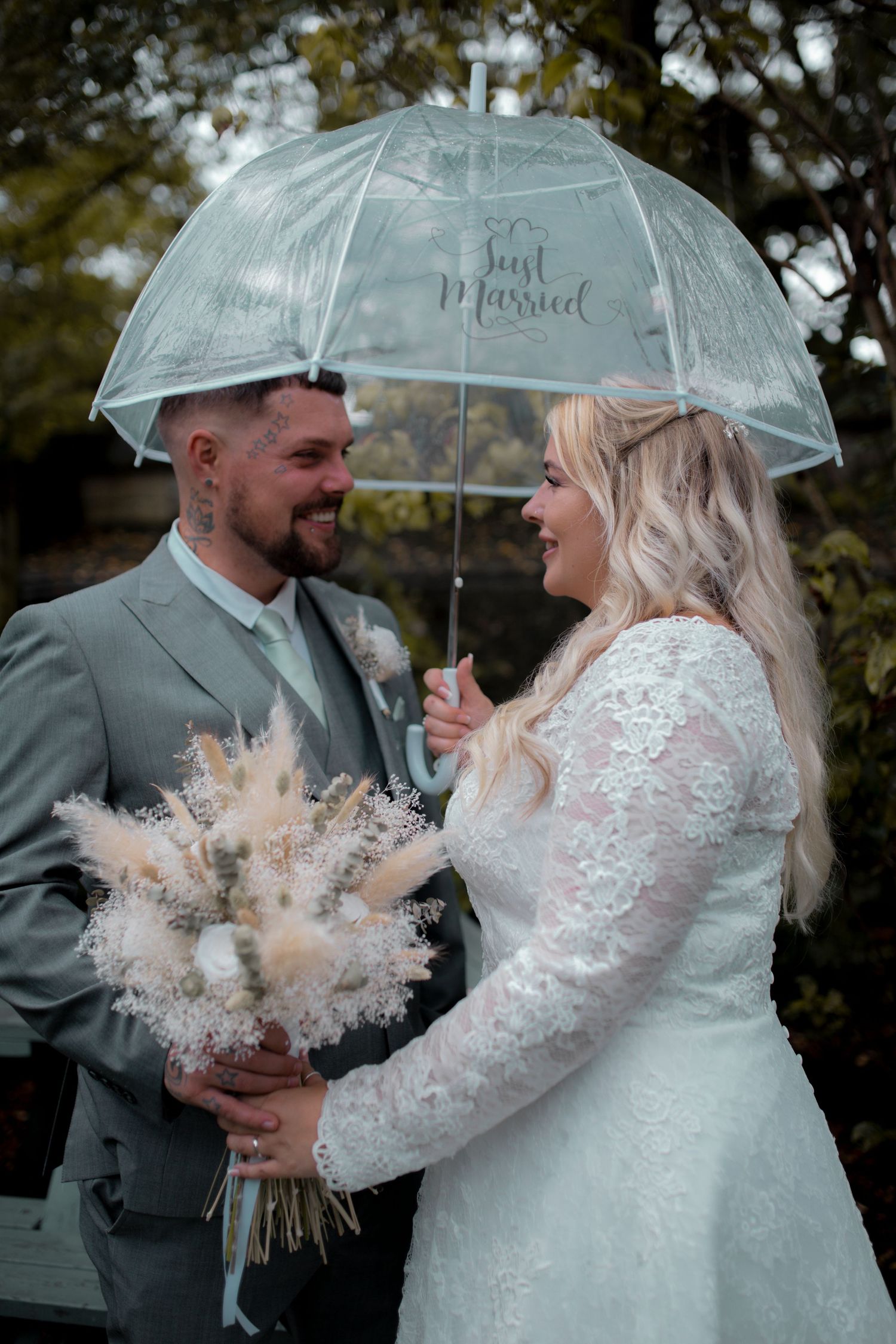 Newlyweds share tender moment under clear umbrella with dried flower bouquet.