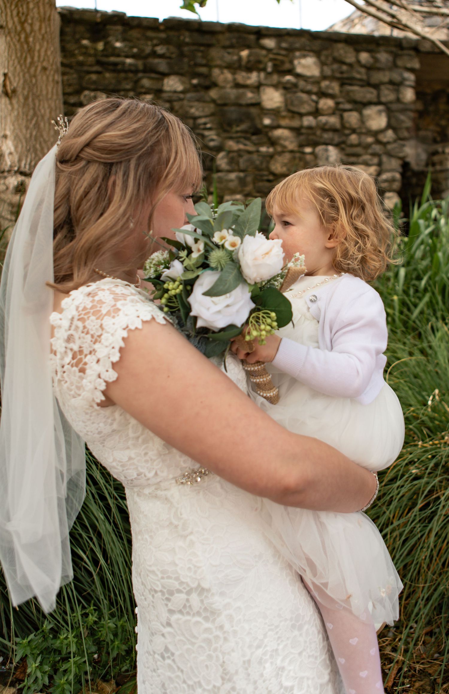 Bride in lace dress shares tender moment with young flower girl against stone wall backdrop.
