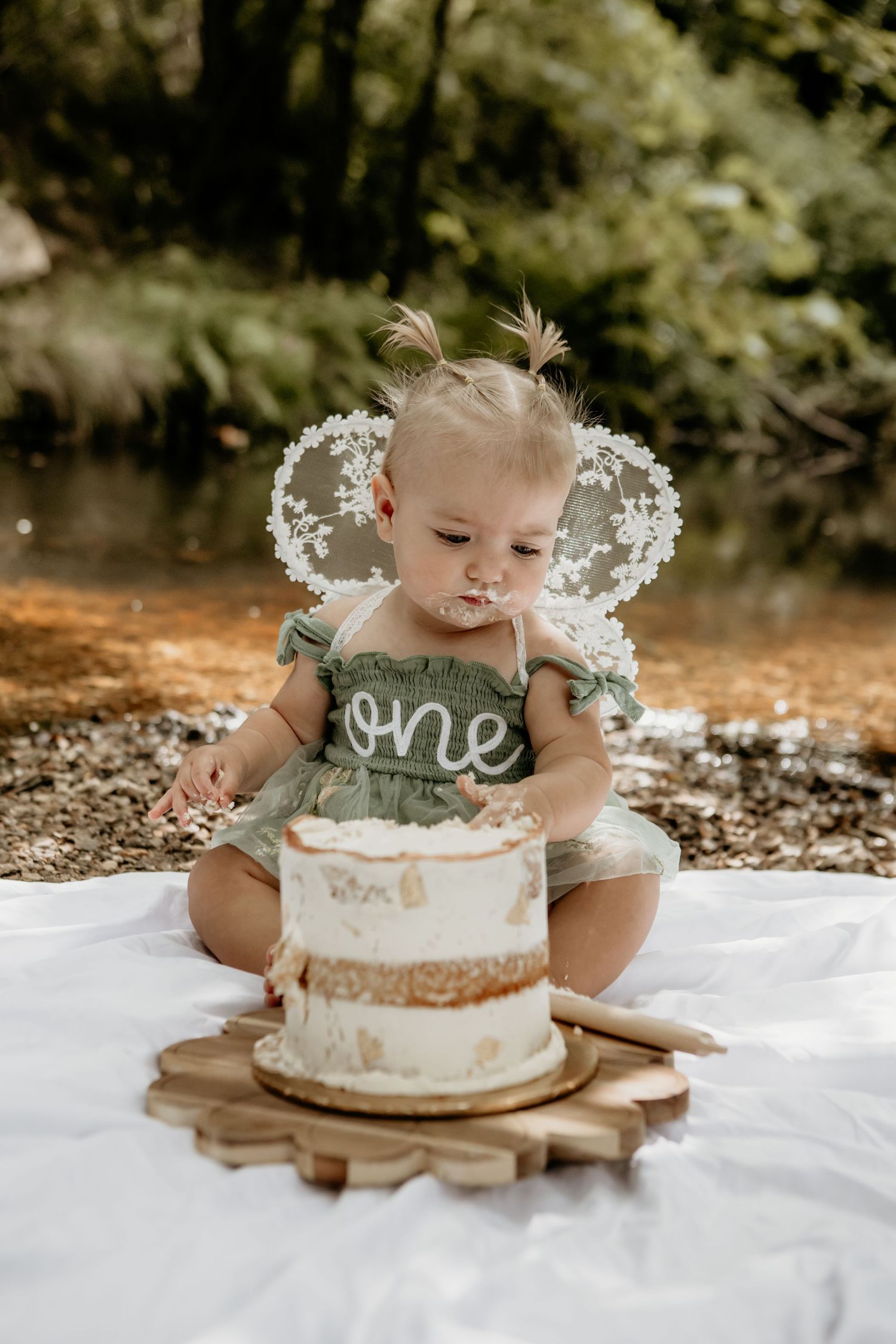 White cake with delicate floral decorations sits on wooden platter during outdoor photoshoot.