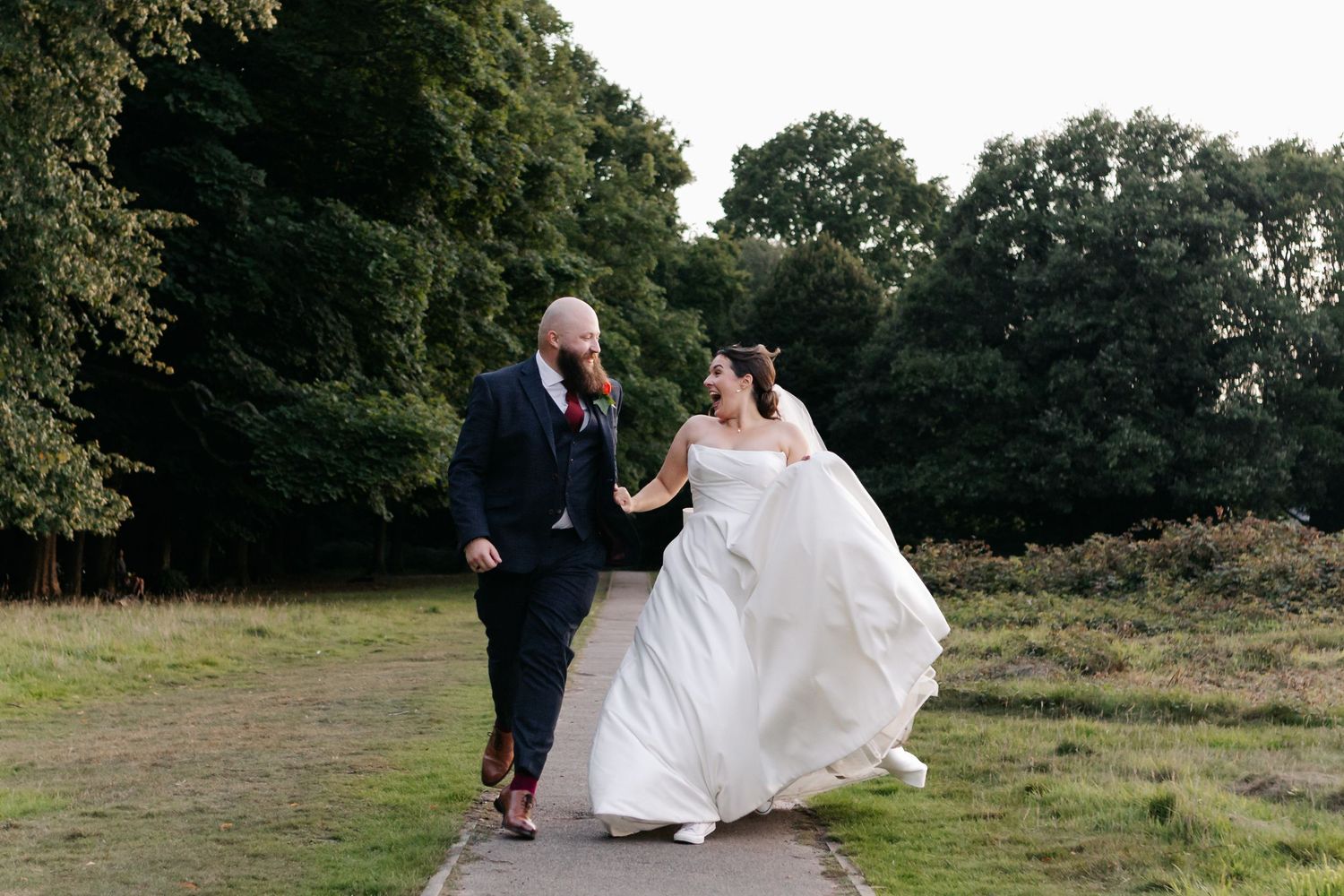Wedding couple dancing playfully on a grassy field surrounded by trees.