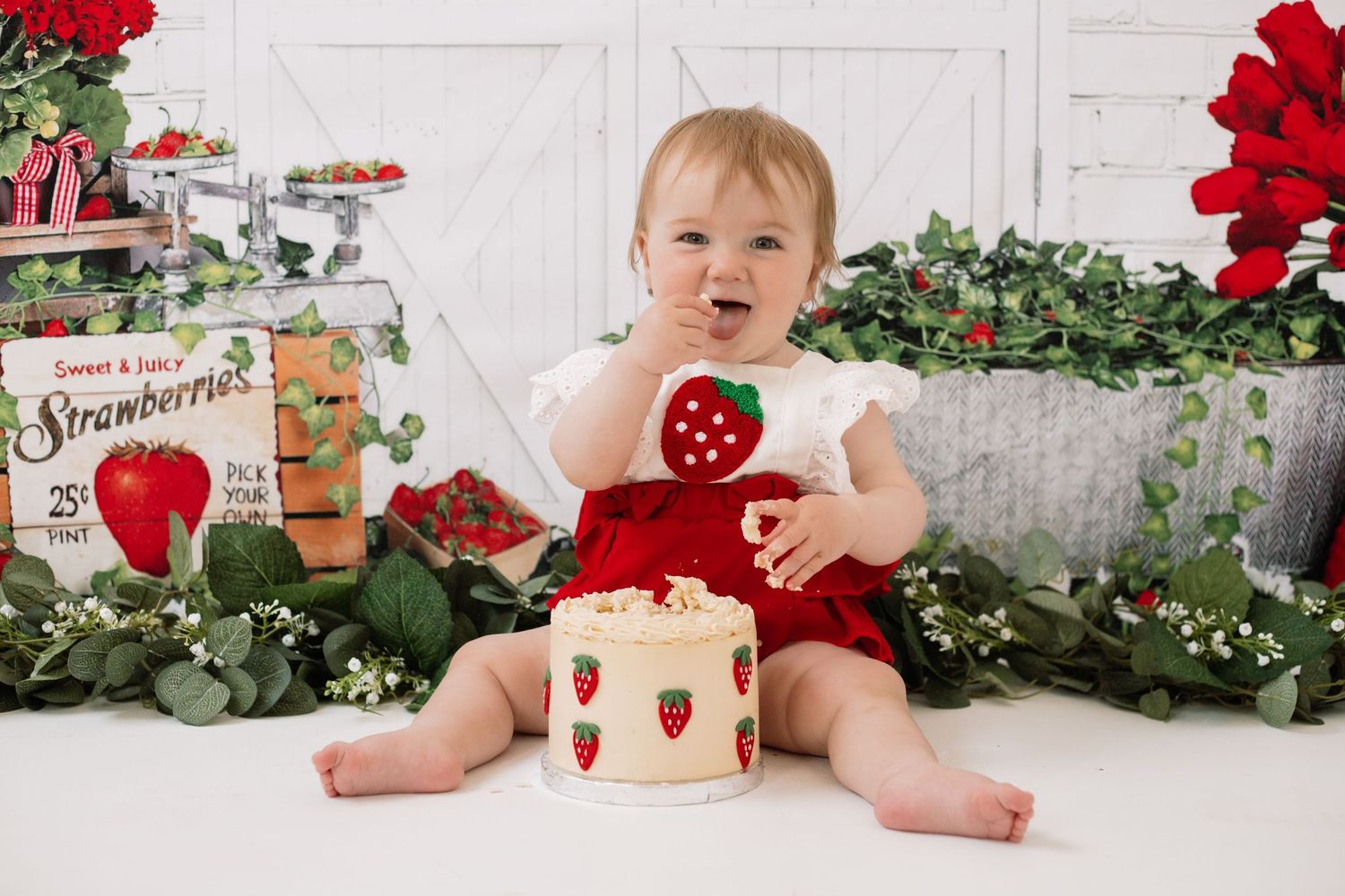Baby sitting on floor with strawberry decorations and red floral accents in studio setting.