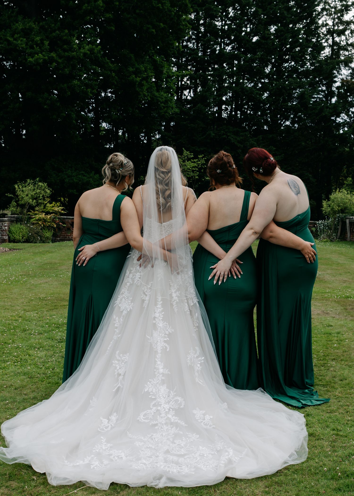 Bridesmaids in emerald green dresses embrace on a lawn with the bride in a flowing white gown.