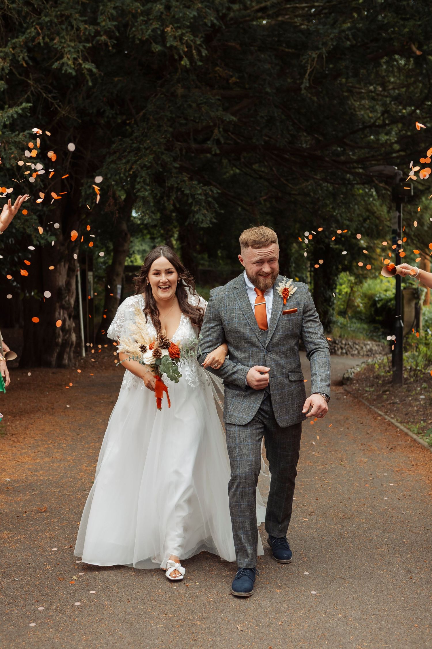 Newlyweds walk together on path surrounded by trees with string lights in background.
