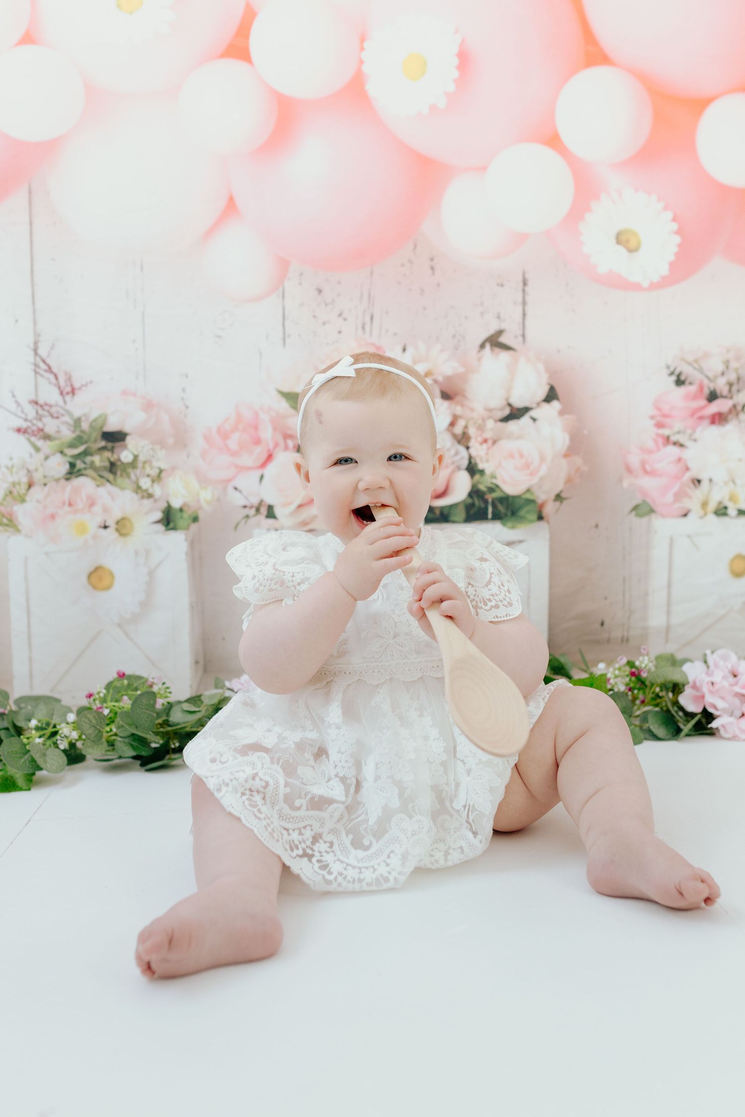 Baby in white lace dress sitting among pink balloons and floral decorations.