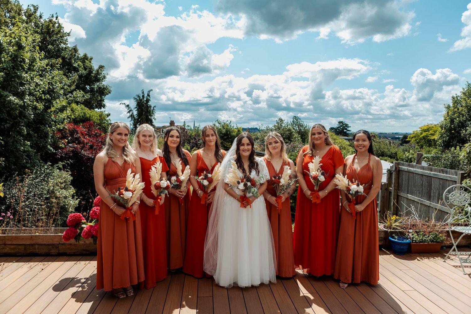 Wedding party in red bridesmaid dresses and white gown pose together outdoors under blue sky.