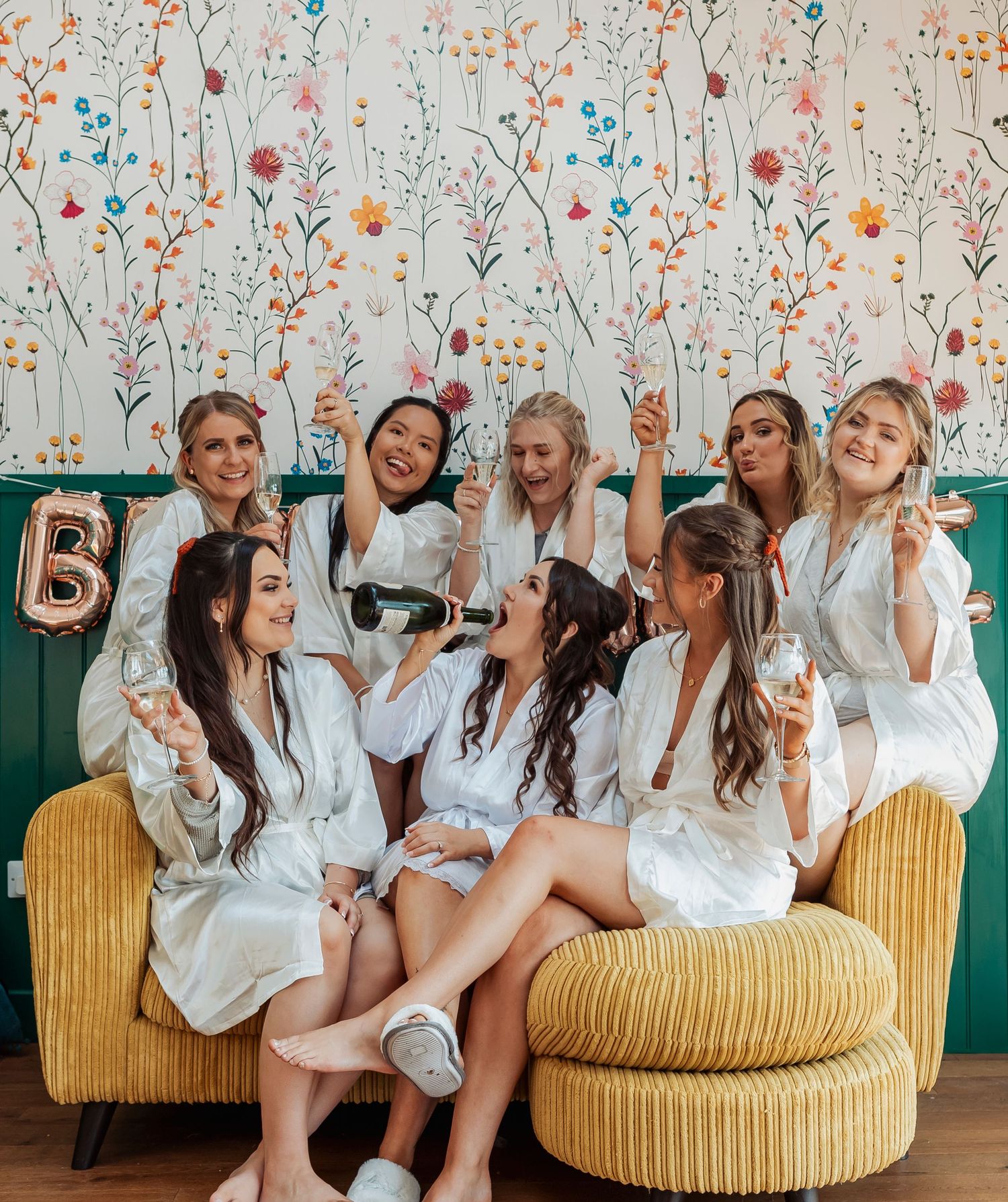 Bridal party in matching white robes laughing together on a mustard yellow couch.