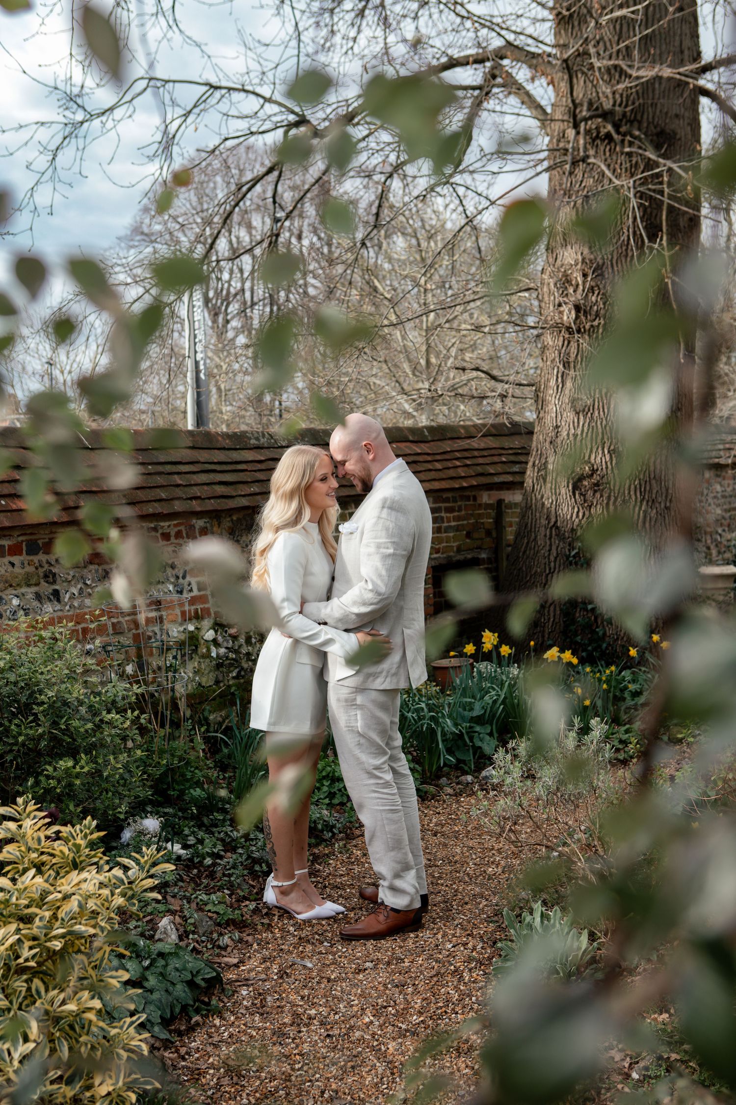 Romantic moment captured in a garden setting with daffodils and brick wall backdrop.
