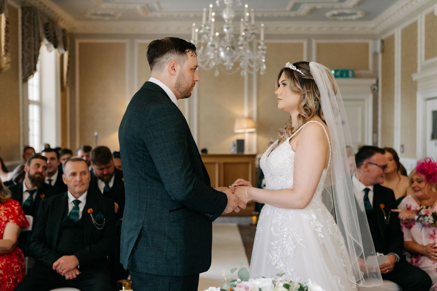 Elegant wedding ceremony taking place in a grand room with ornate chandeliers and white decor.