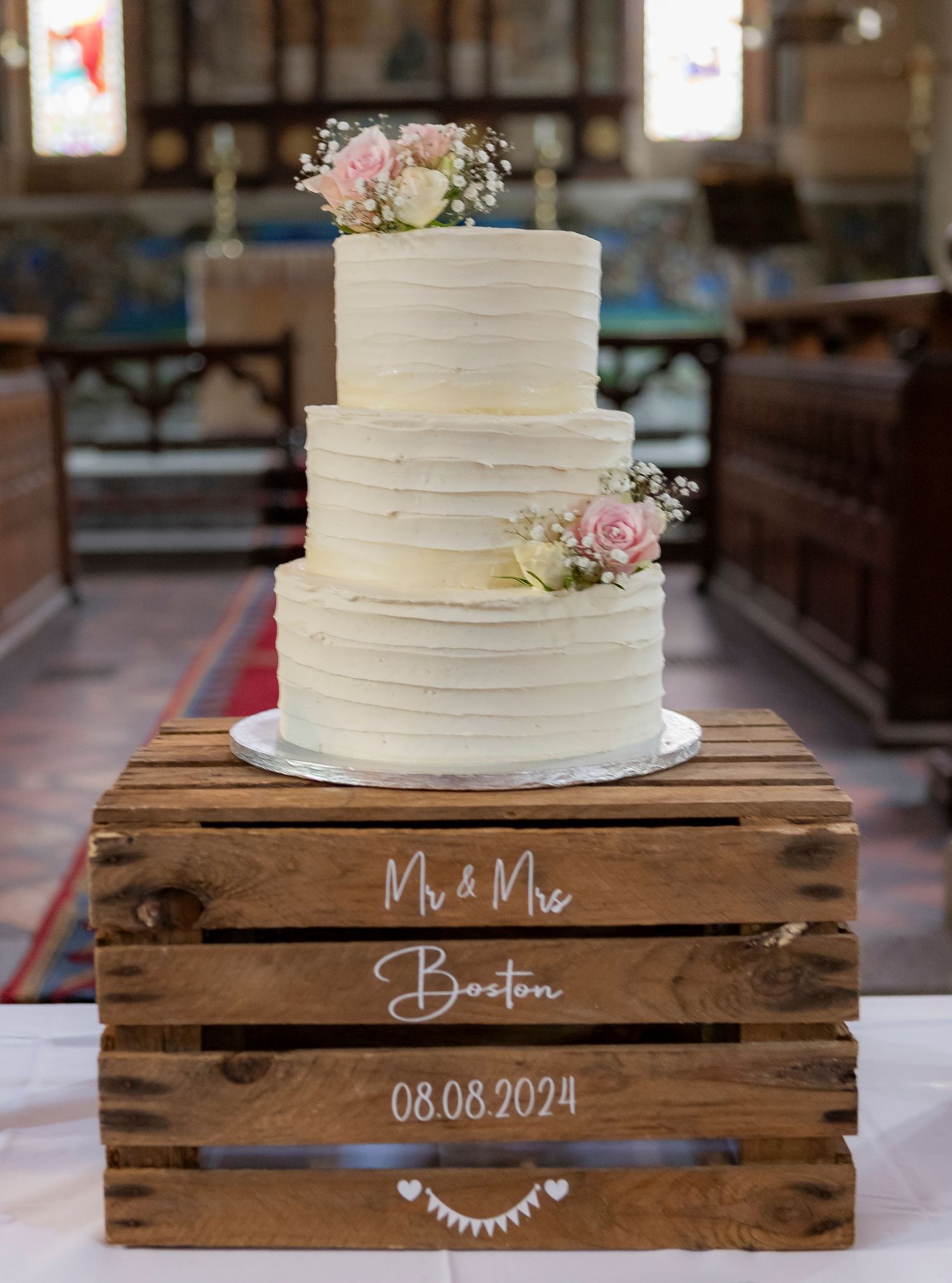 White tiered wedding cake displayed on rustic wooden crates with delicate floral decorations.