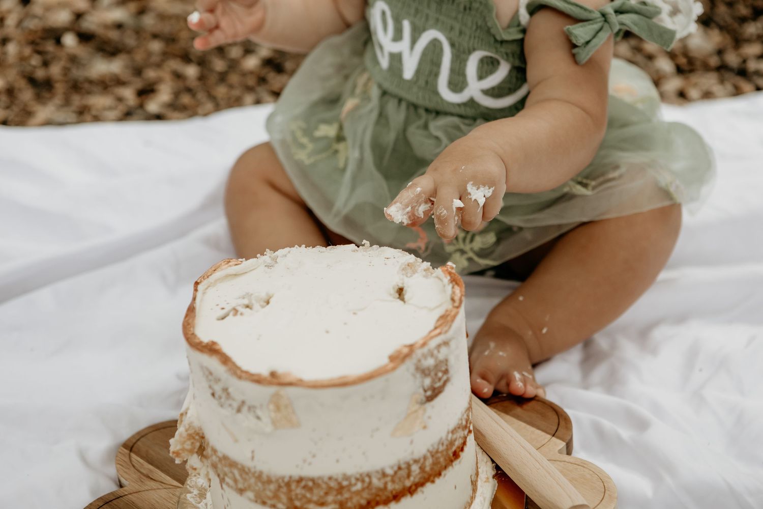 Baby in sage green outfit reaches for white cake during outdoor photo session.