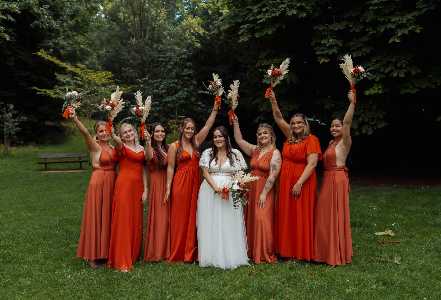 Wedding party in red dresses poses on grass with white-gowned bride.
