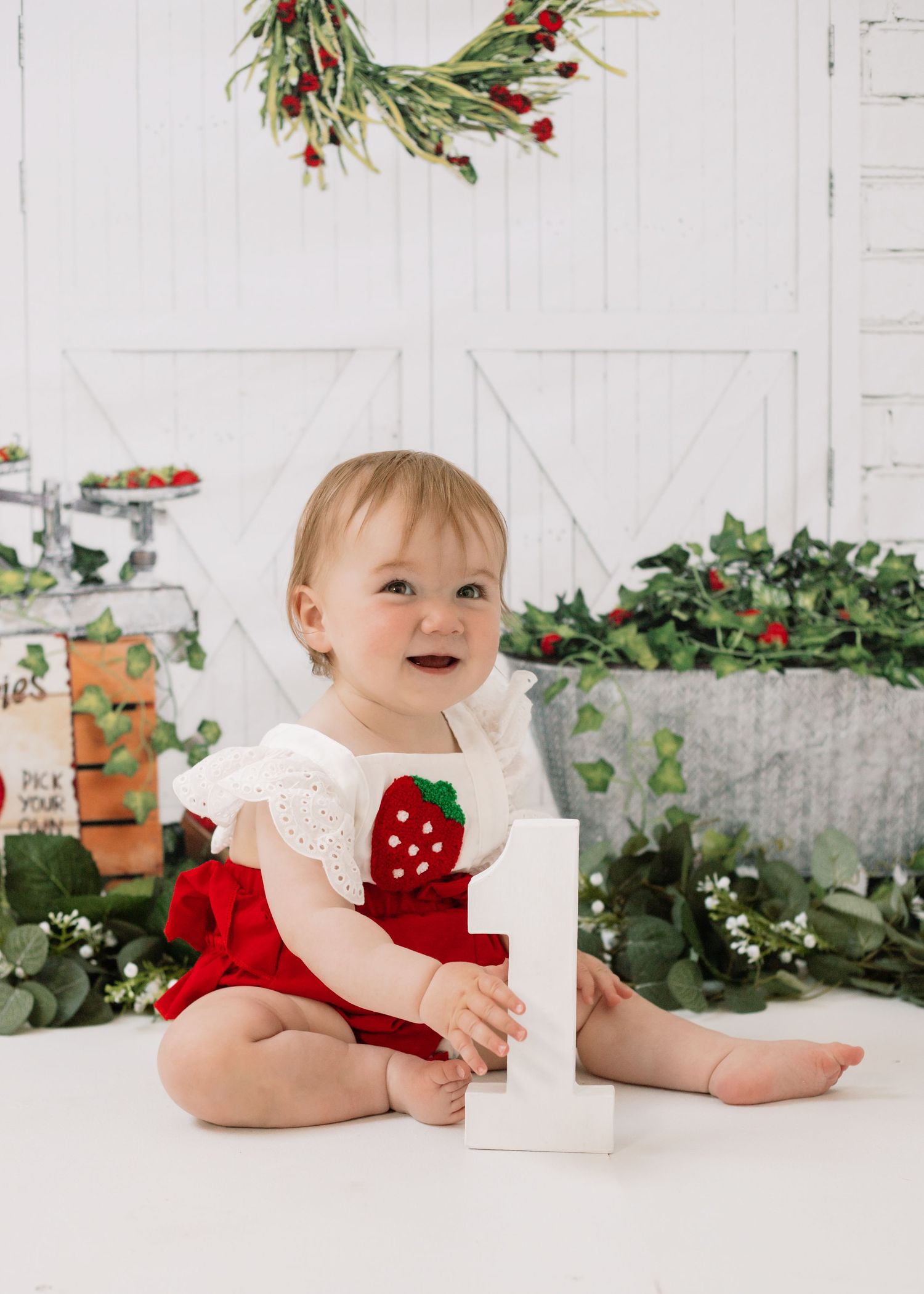 Baby in red and white strawberry-themed outfit sits on white floor with greenery and holiday decor.