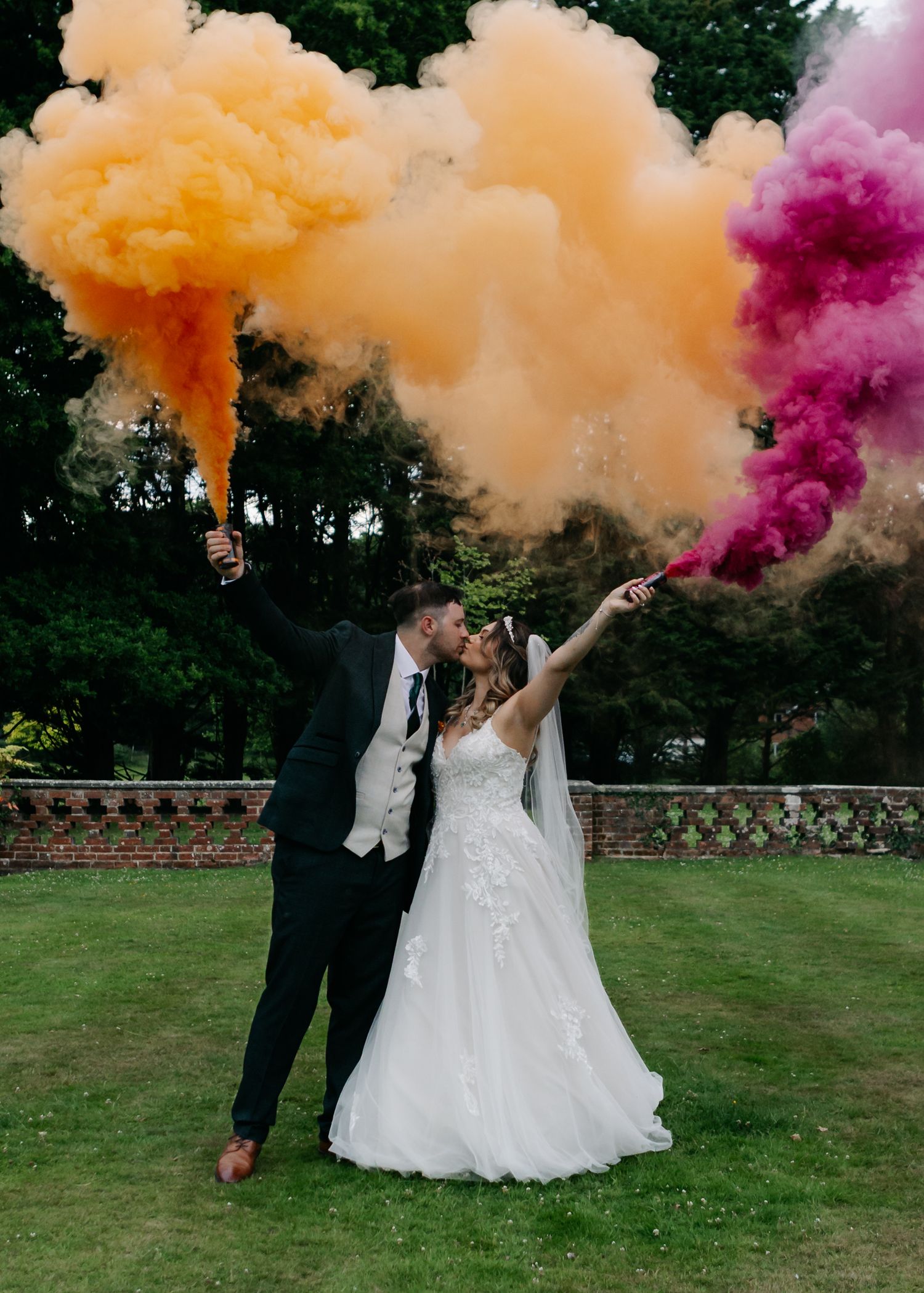 Wedding couple celebrates with colorful smoke bombs in orange and pink on a grassy lawn.