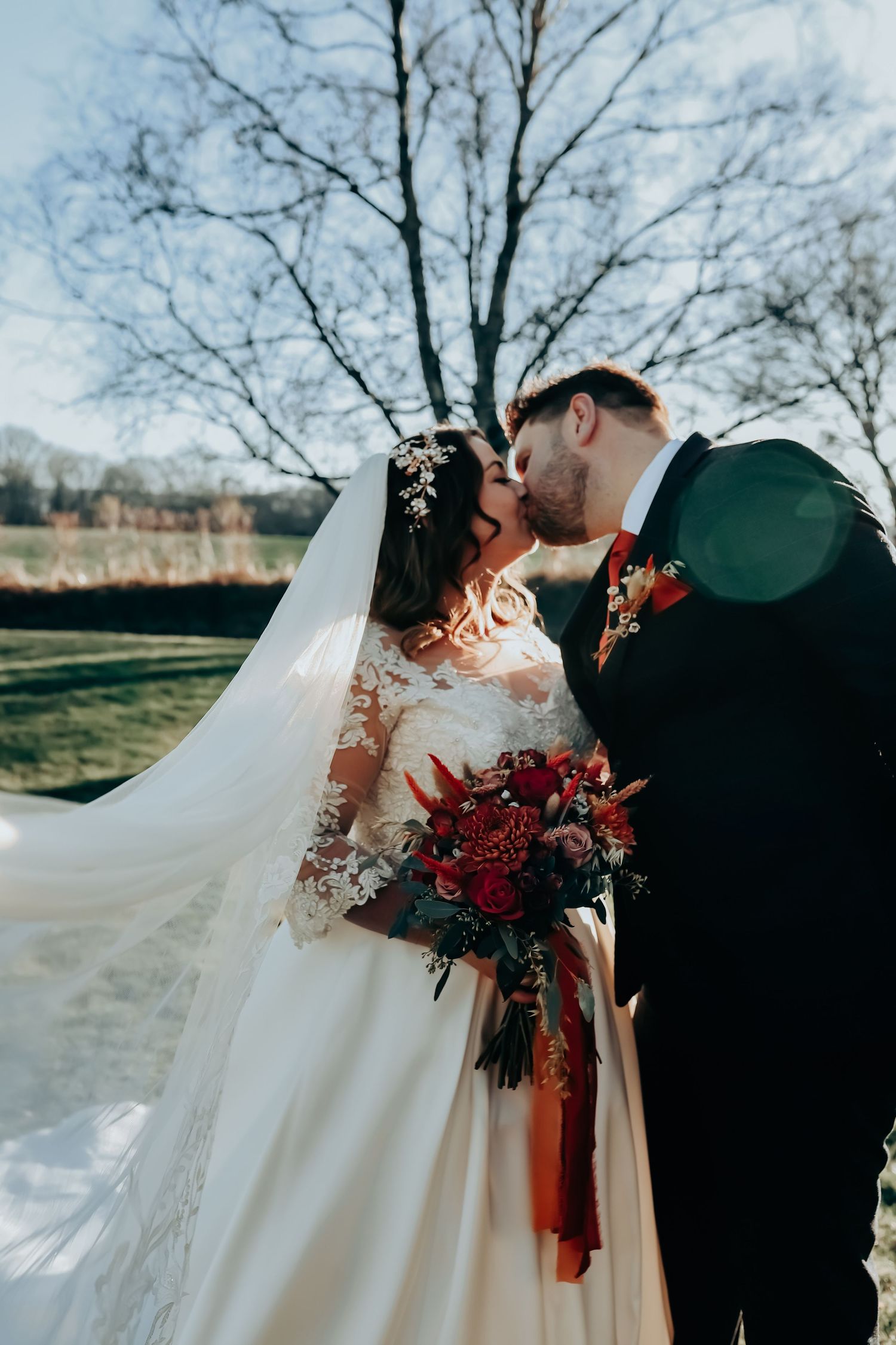 Romantic winter wedding photo with flowing white dress and red bouquet against bare trees.